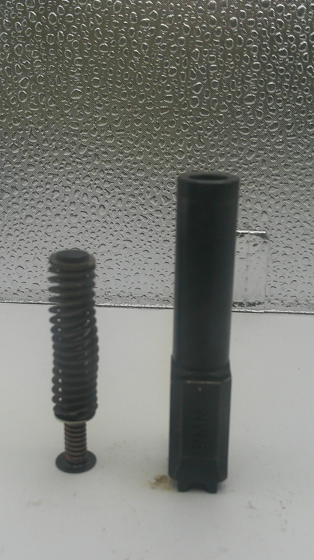 A black metal wall-mounted mounting bracket with a long threaded screw, shown against a split background of plain white and textured gray.