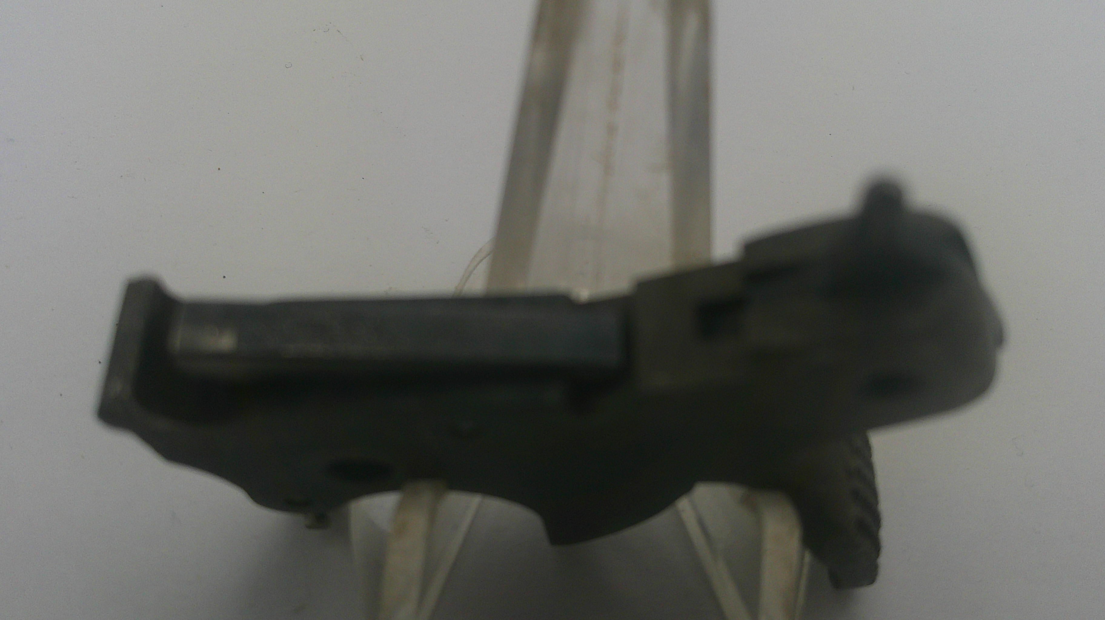 A small black plastic or metal component photographed against a plain light background, held by a clear plastic stand.