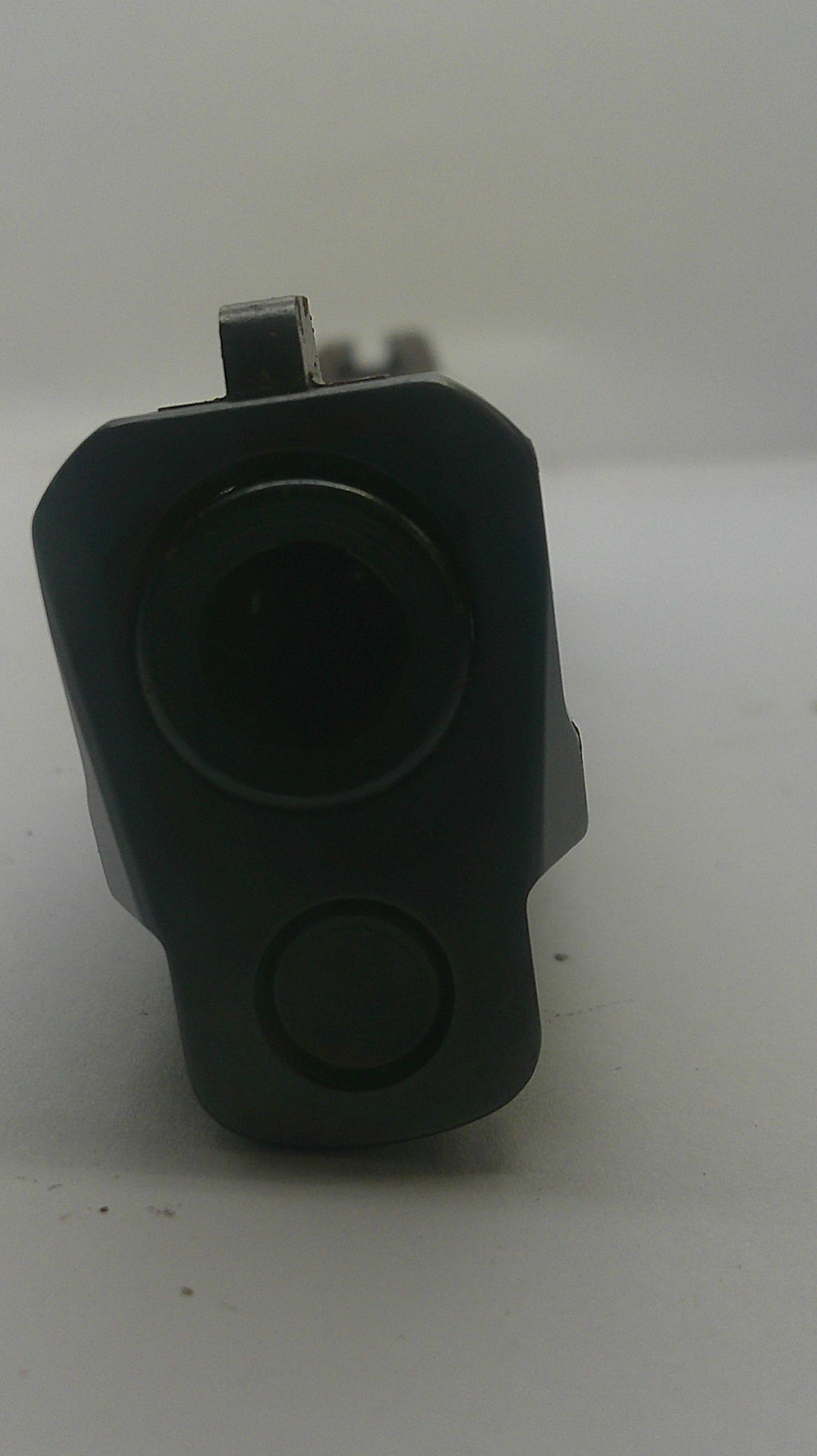 A black plastic automotive camera lens module with a mounting tab, photographed on a plain light gray surface.