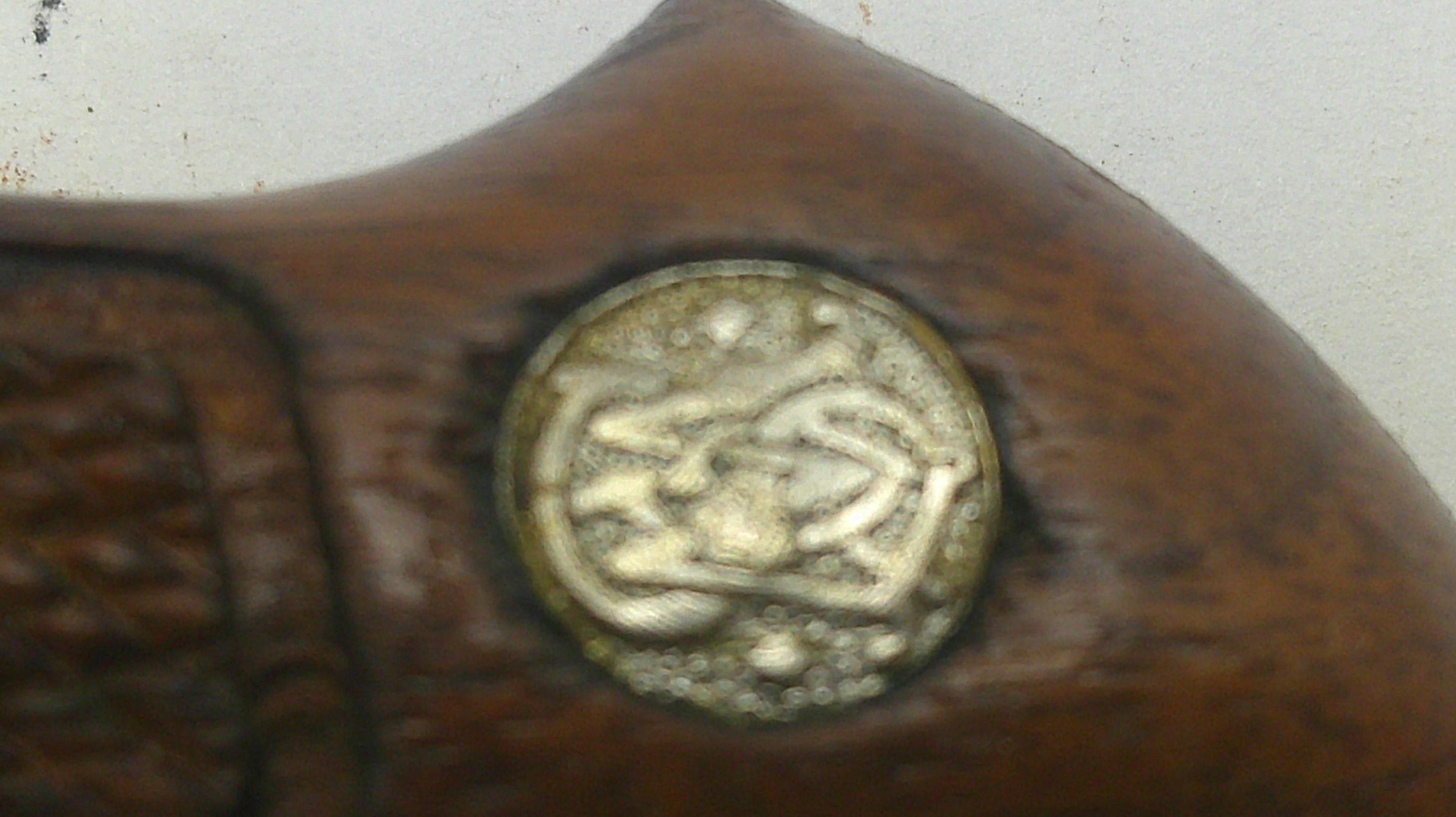 Close-up of a dark brown wooden object with a round metallic emblem featuring Arabic calligraphy, set against a light off-white background.