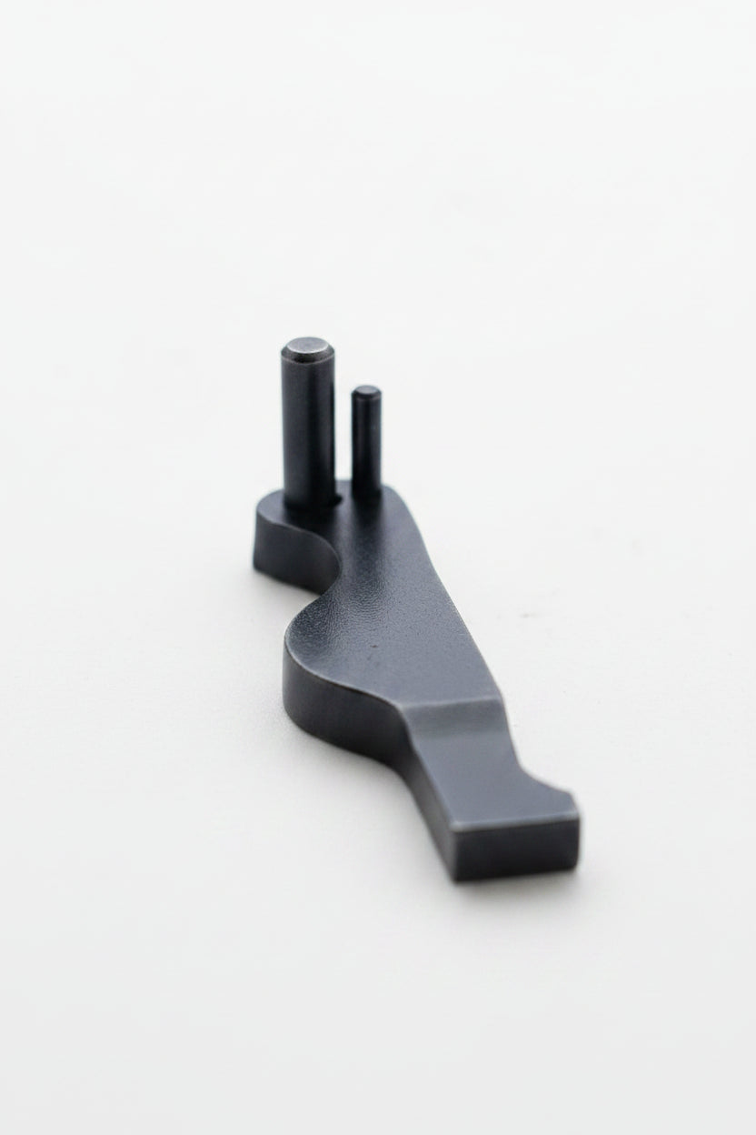 A small dark metal mechanical part with two protruding pins, shown against a plain off-white background.
