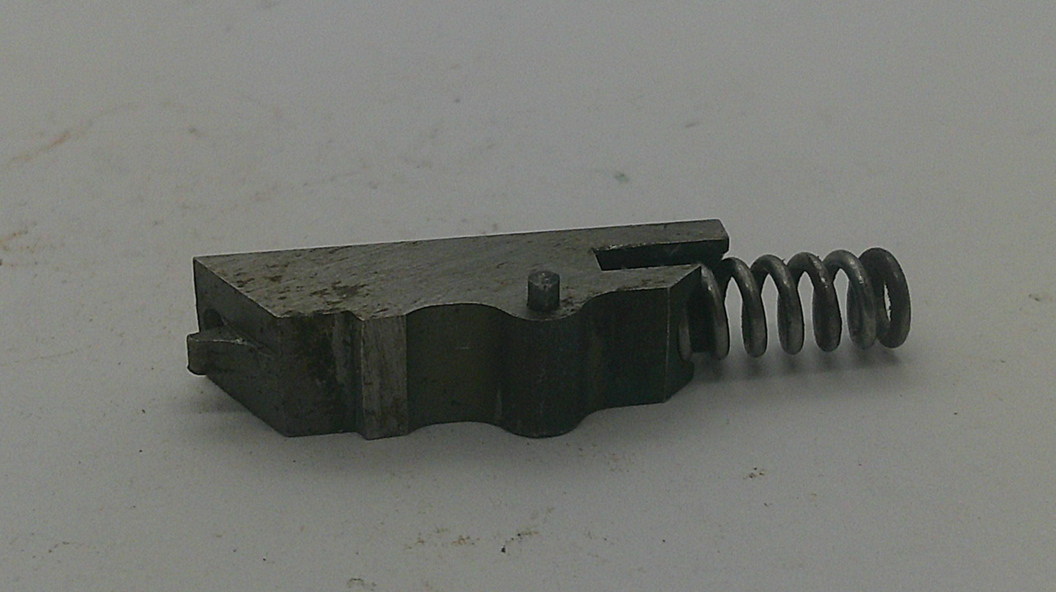 A small dark metal mechanical component with a coiled spring attached, photographed on a plain off-white surface.