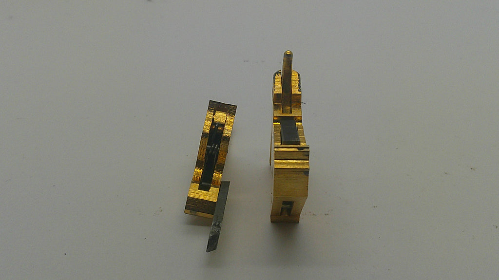 Two small gold-toned metal hardware components with dark internal details, photographed on a plain light gray surface.