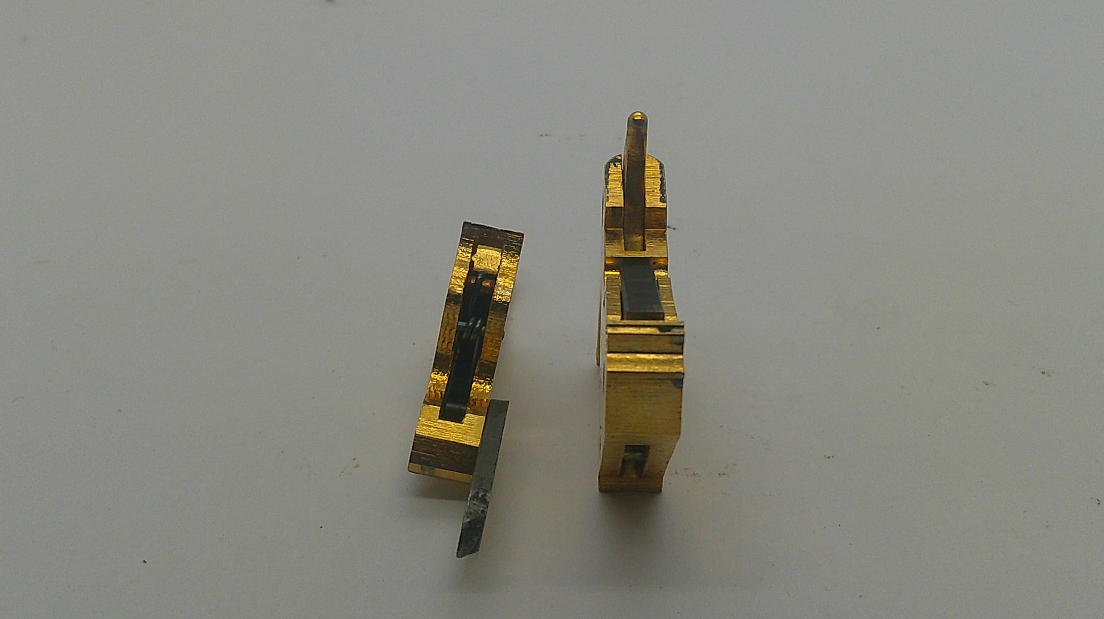 Two small gold-toned metal hardware components with dark internal details, photographed on a plain light gray surface.