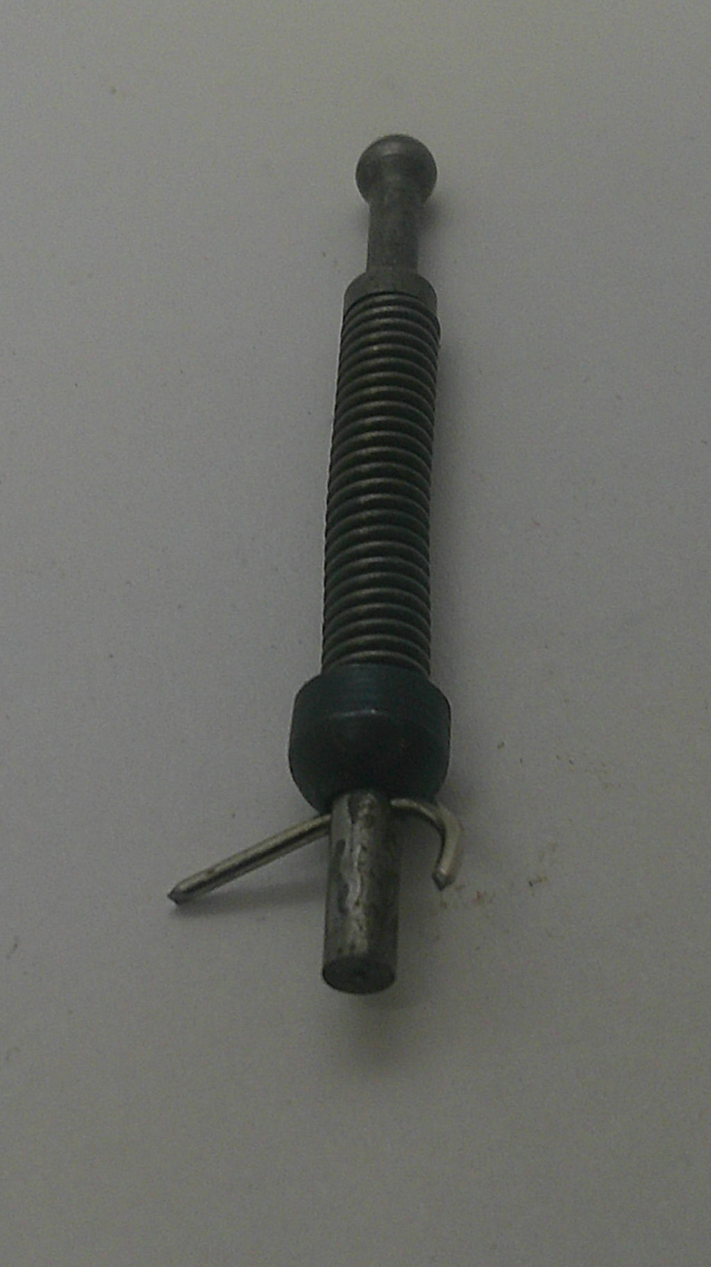 A small metal mechanical component with a threaded rod, a dark green plastic or rubber ring, and a bent metal pin, photographed on a plain light gray surface.