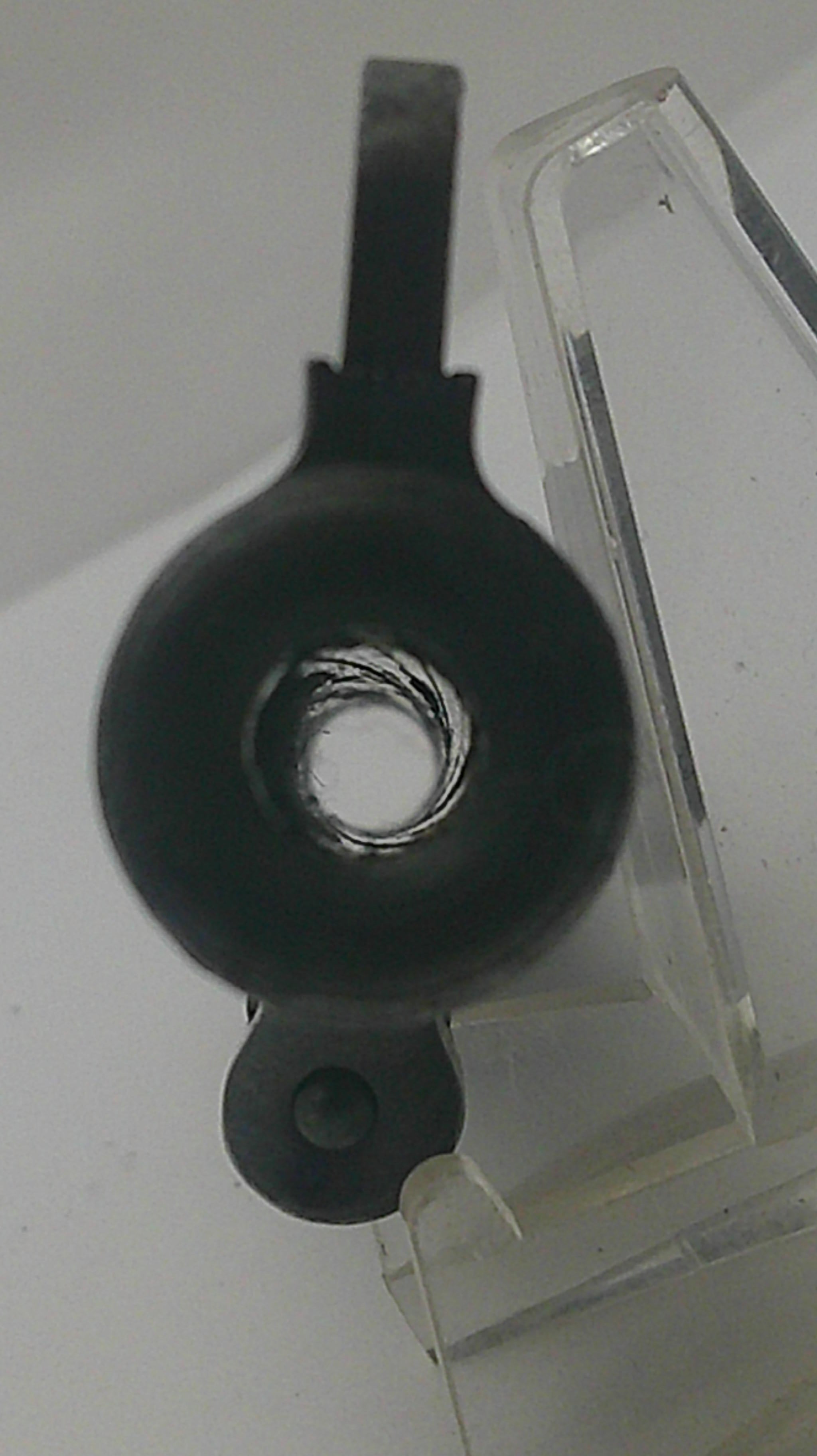 A close-up of a black circular ring-shaped object with a central hole and a flat tab extending to the right, resting on a clear acrylic stand against a light gray background.