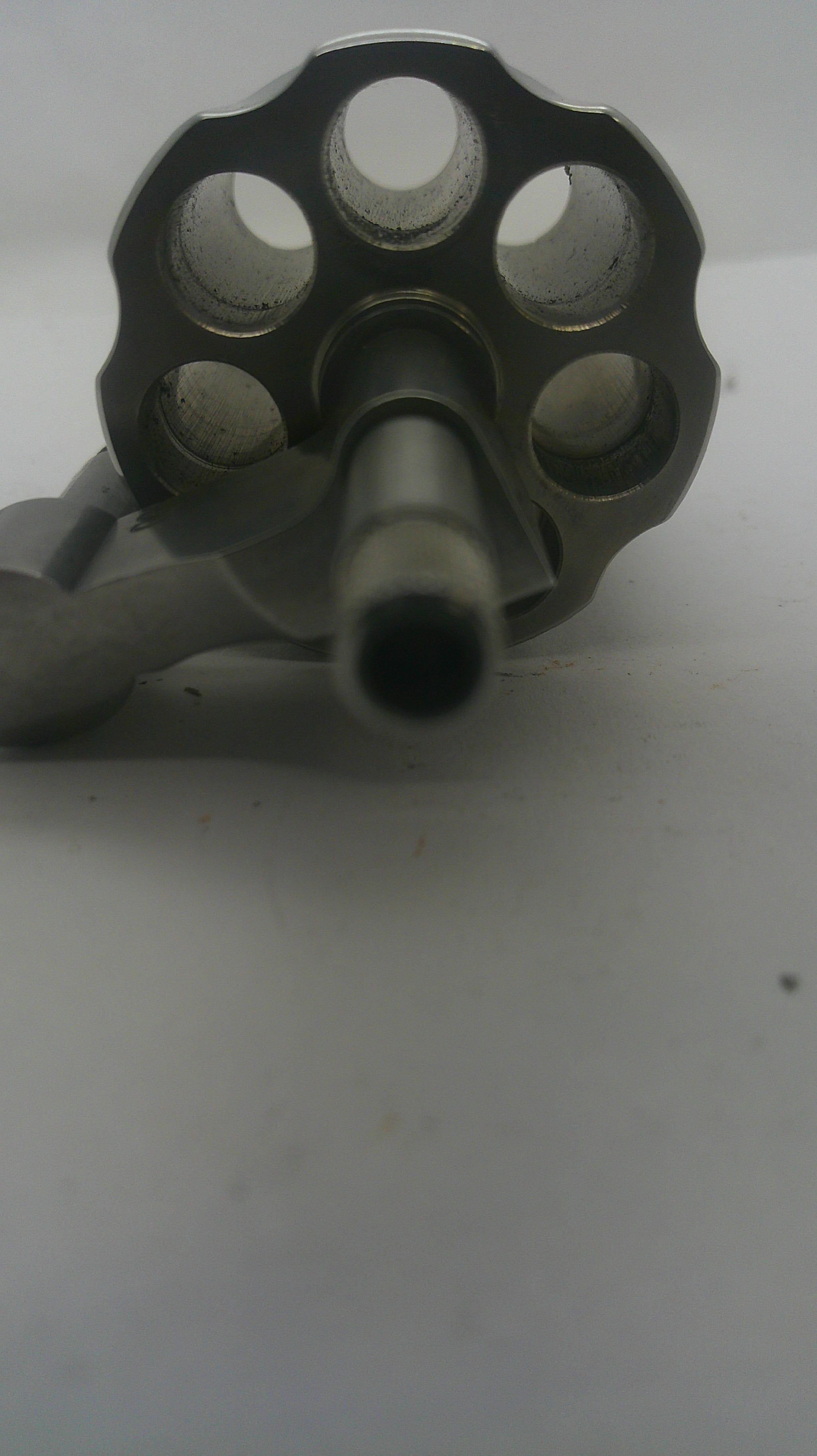A close-up shot of a metallic revolver-style object with a multi-hole circular end and a central barrel, photographed against a plain light background.