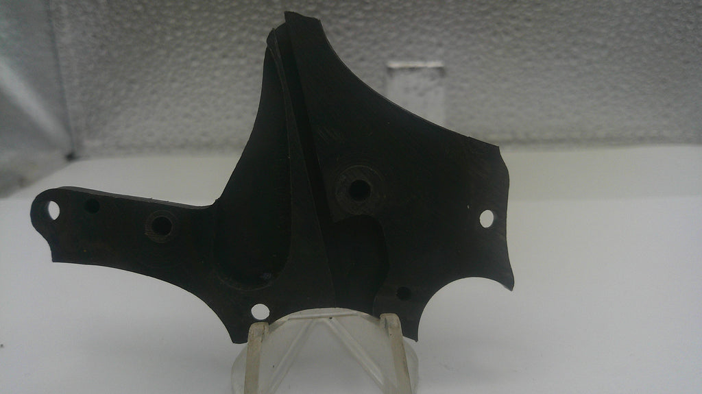 A black plastic engine or mechanical bracket with mounting holes, displayed on a clear plastic stand against a light gray textured background.