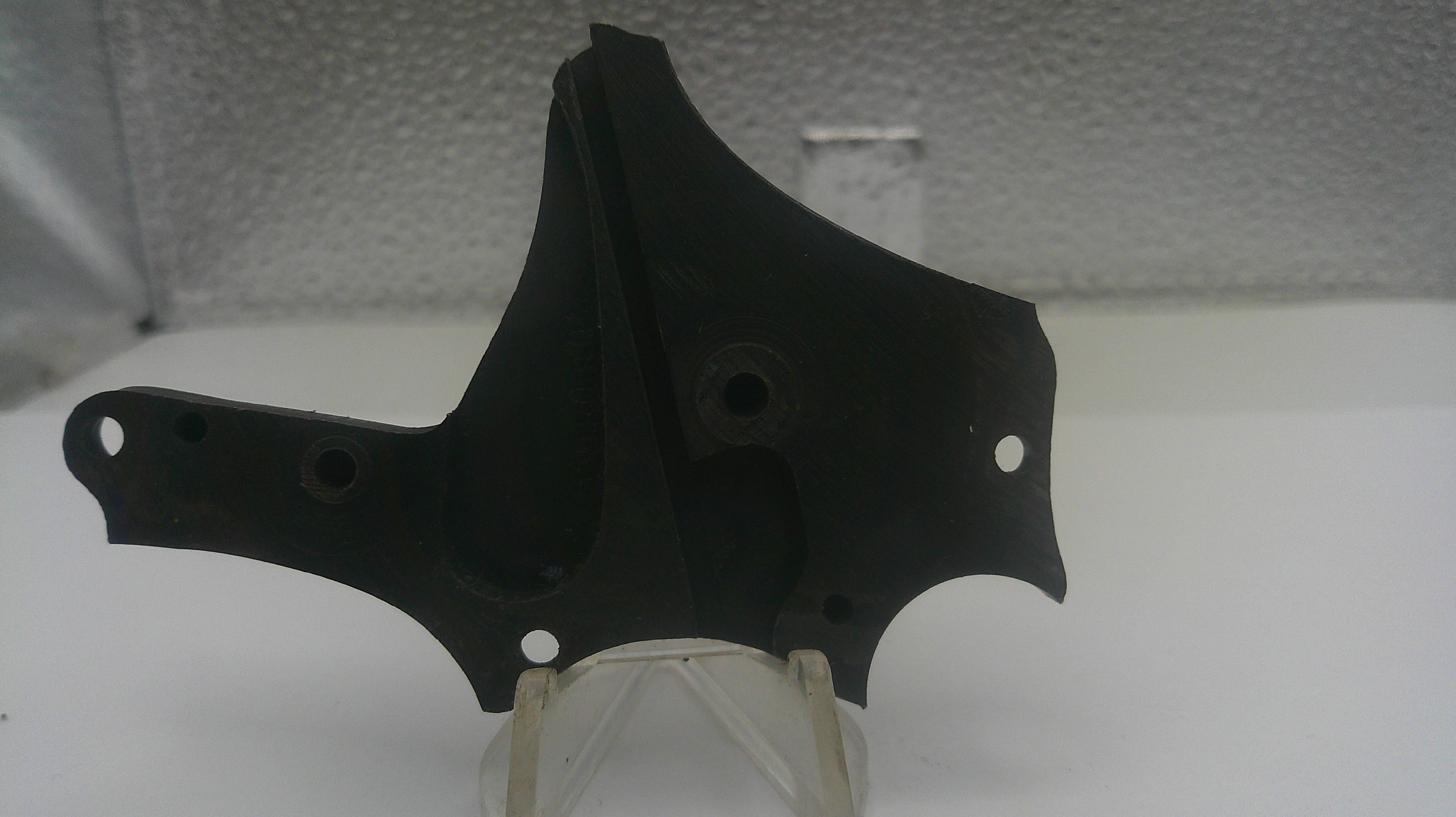 A black plastic engine or mechanical bracket with mounting holes, displayed on a clear plastic stand against a light gray textured background.