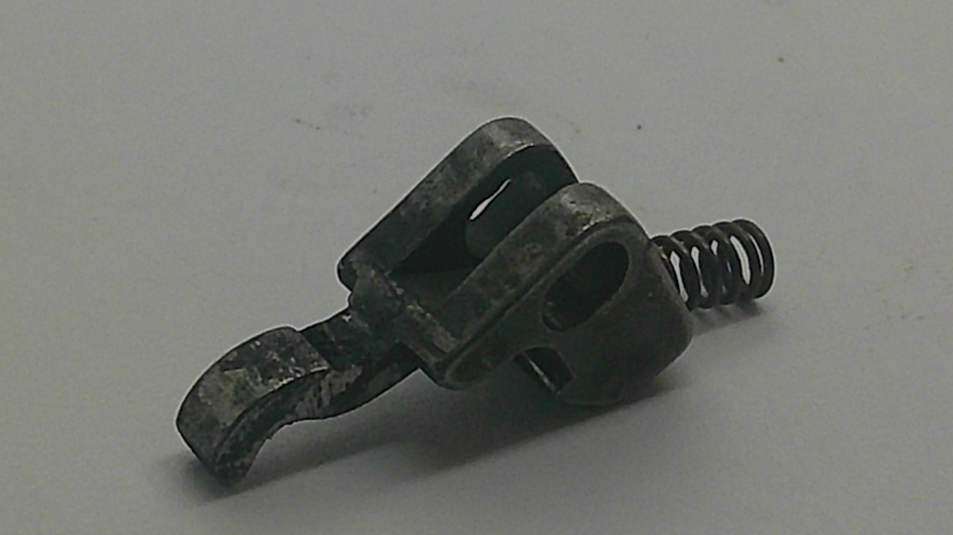 A small dark metal hardware component with a hook-like arm and a coiled spring, photographed on a plain light gray background.