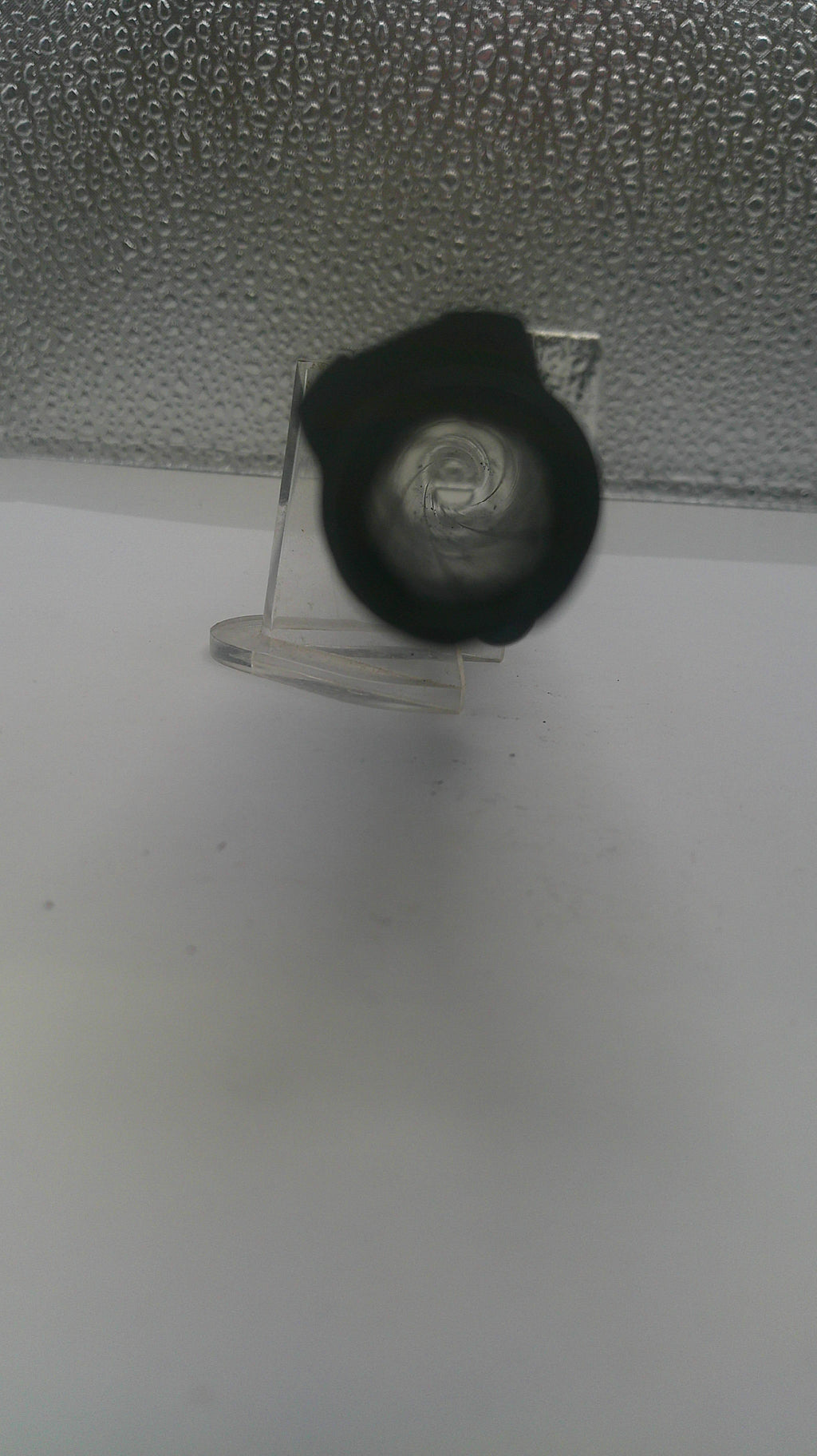A close-up of a black circular object, possibly a lens or filter, mounted on a clear plastic stand against a textured gray background.