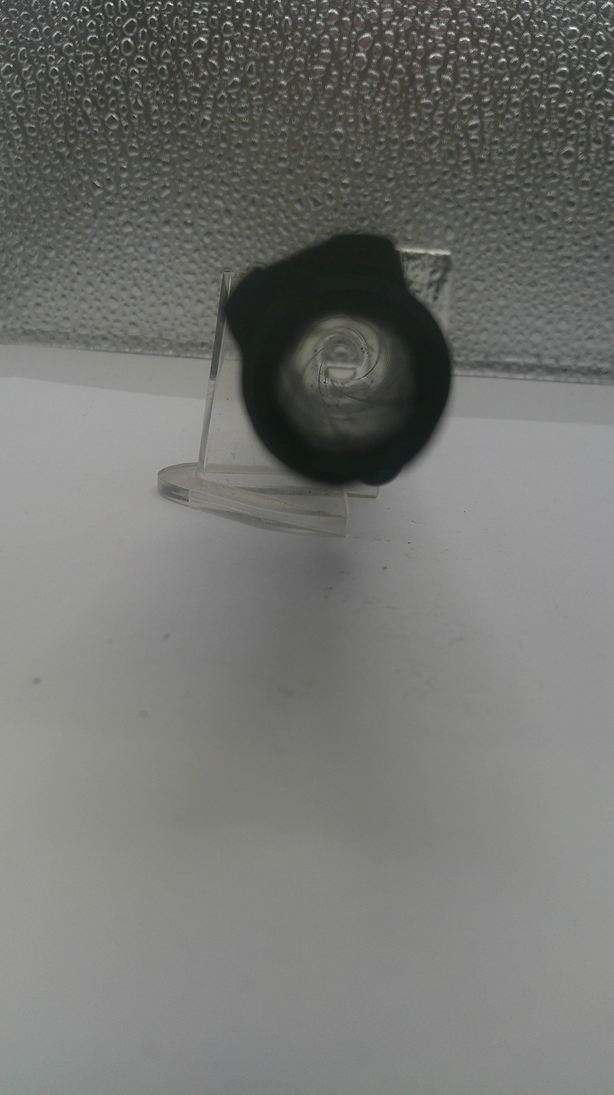 A close-up of a black circular object, possibly a lens or filter, mounted on a clear plastic stand against a textured gray background.