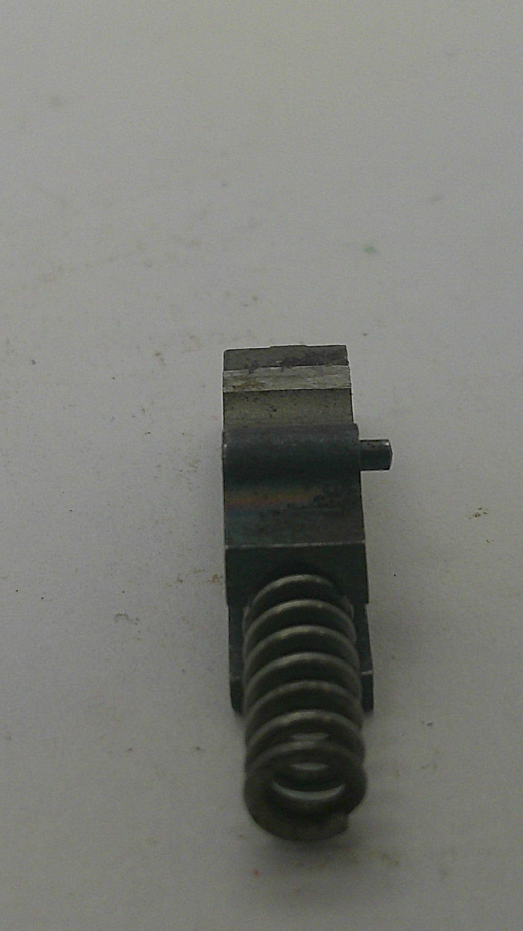 A small dark metal mechanical component with a coiled spring on one end, photographed on a plain off-white surface.