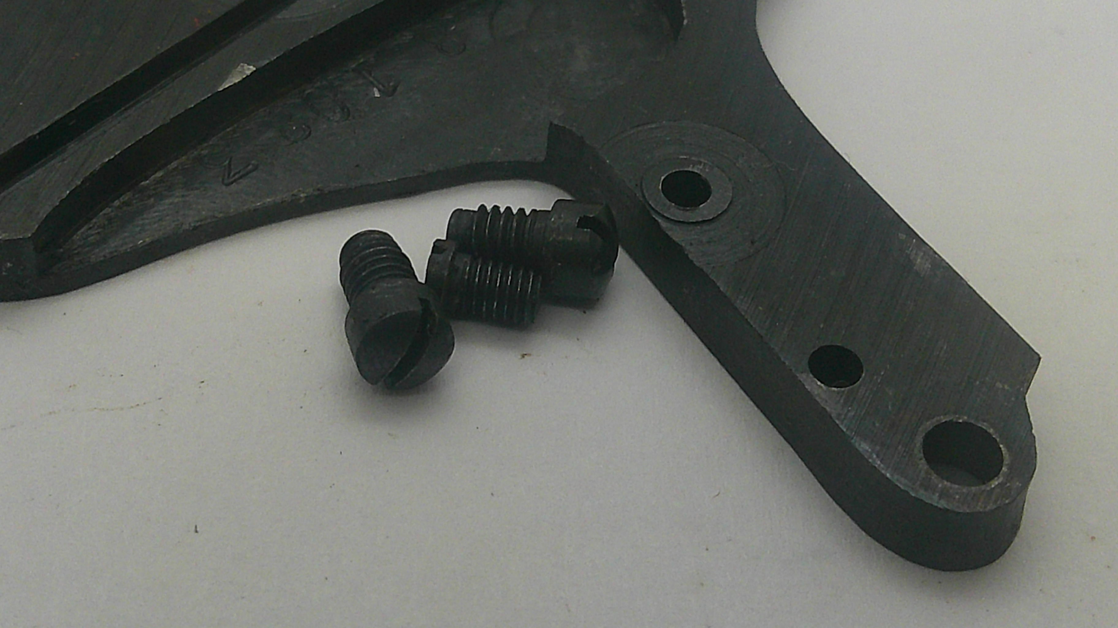 A close-up of a black metal mechanical component with three small black threaded fasteners placed on a light gray surface.
