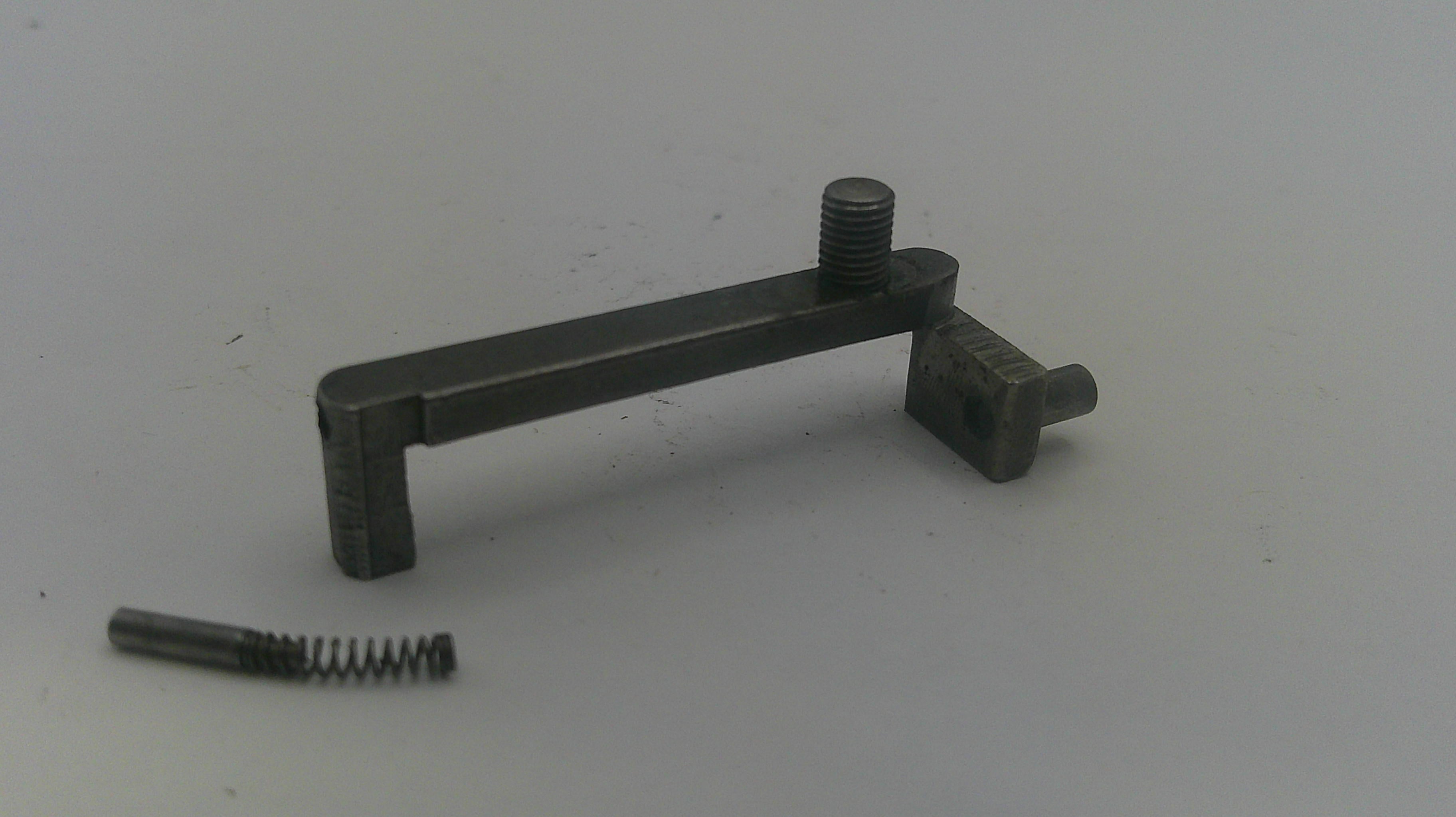 A small dark metal mechanical component with a threaded rod and a separate small spring, photographed on a plain light gray background.