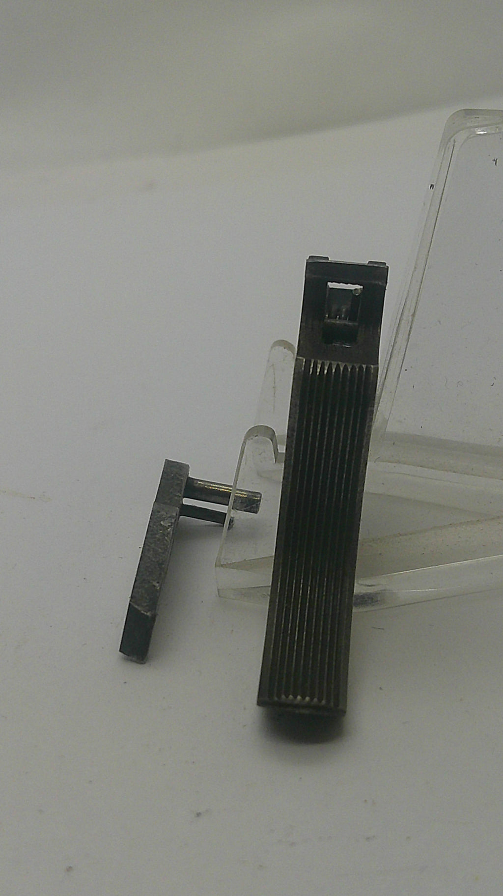A black ribbed rectangular accessory with a small metal clip or hook attached, resting on a clear plastic stand against a plain light background.