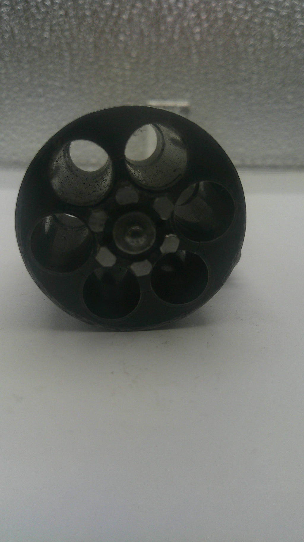 A black plastic six-hole revolver grip or magazine insert, shown lying on a white surface next to a textured gray wall.