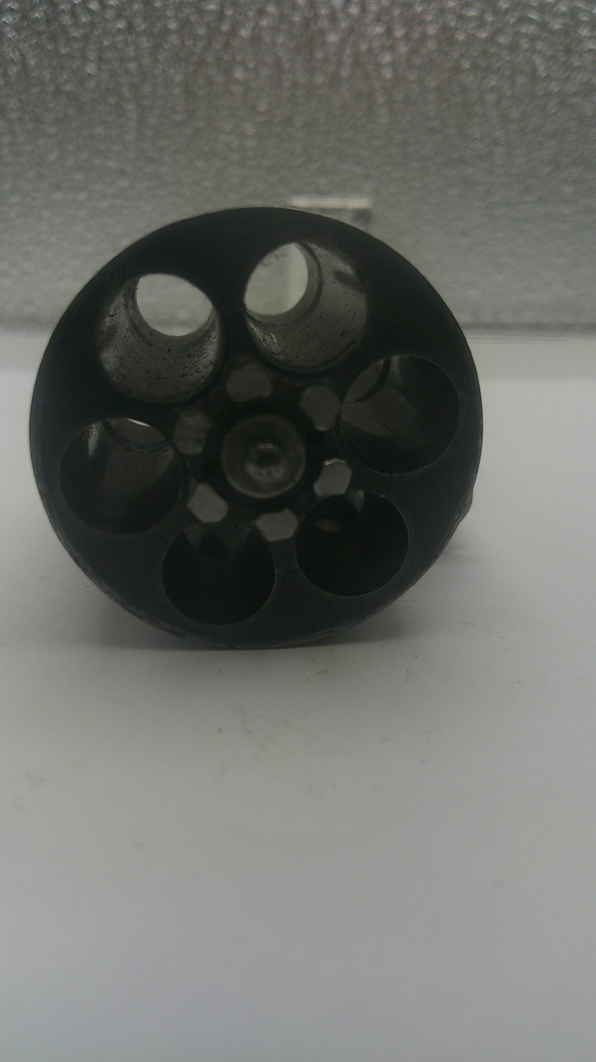 A black plastic six-hole revolver grip or magazine insert, shown lying on a white surface next to a textured gray wall.
