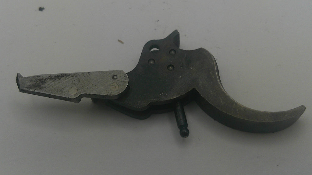 A used metal firearm trigger assembly with a dark gray handle and a weathered silver-toned plate, resting on a plain light gray surface.