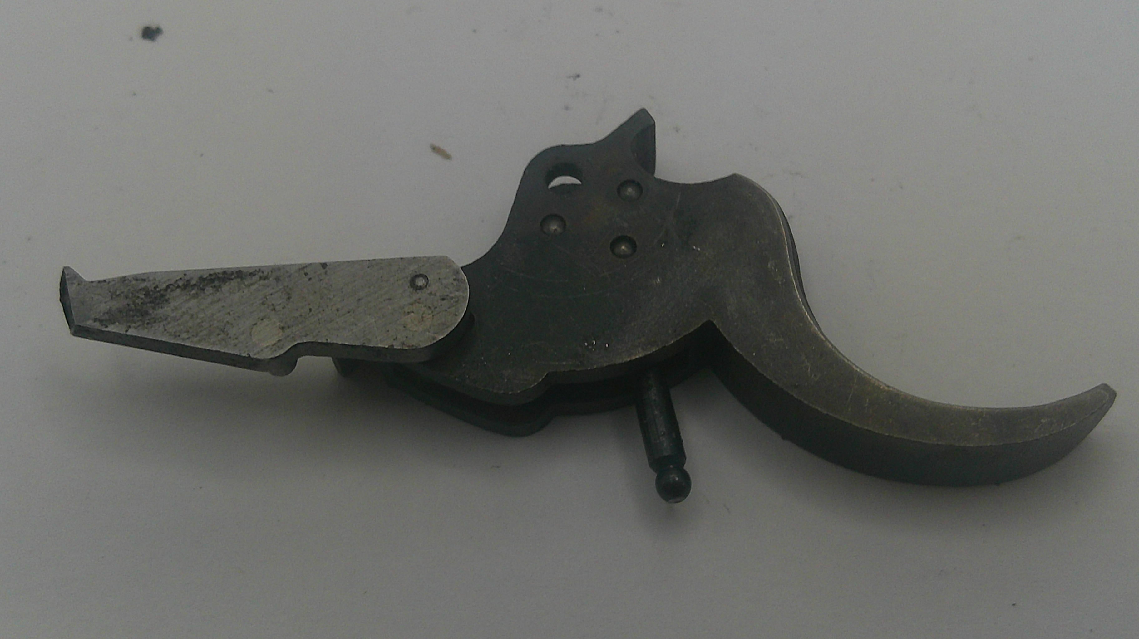 A used metal firearm trigger assembly with a dark gray handle and a weathered silver-toned plate, resting on a plain light gray surface.