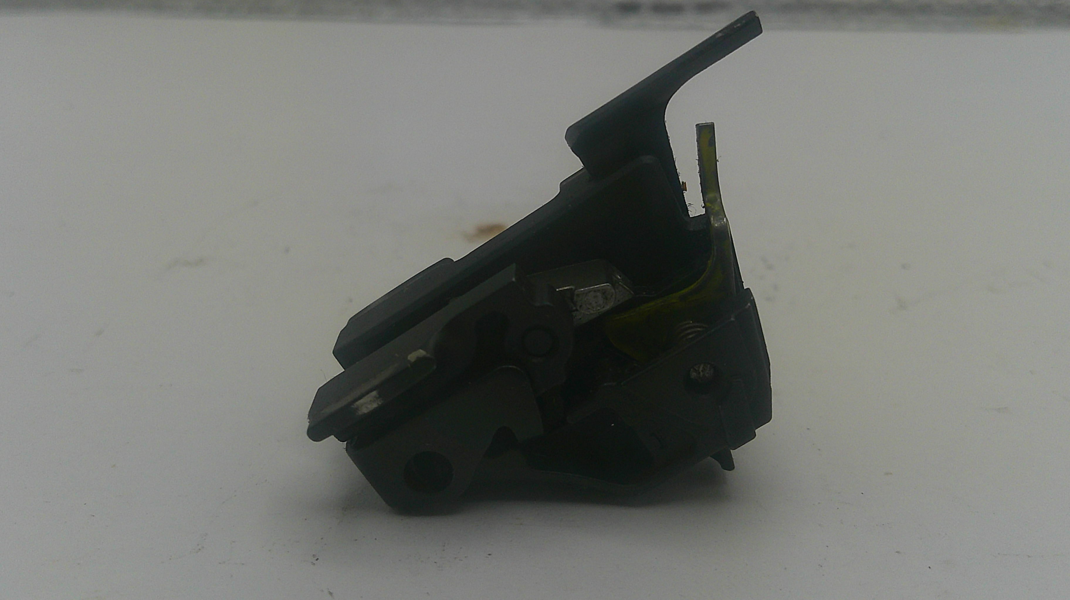 A small black plastic mechanical component or switch assembly with a hinged tab, photographed on a plain light gray surface.