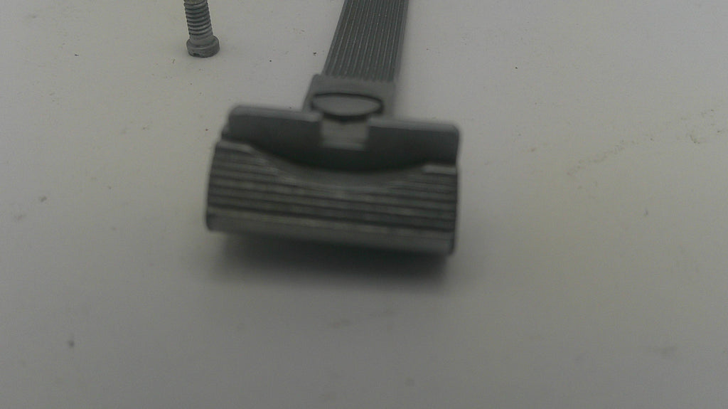 A close-up of a dark gray metal razor head with a textured guard, shown on a plain light gray surface with a small metal screw nearby.