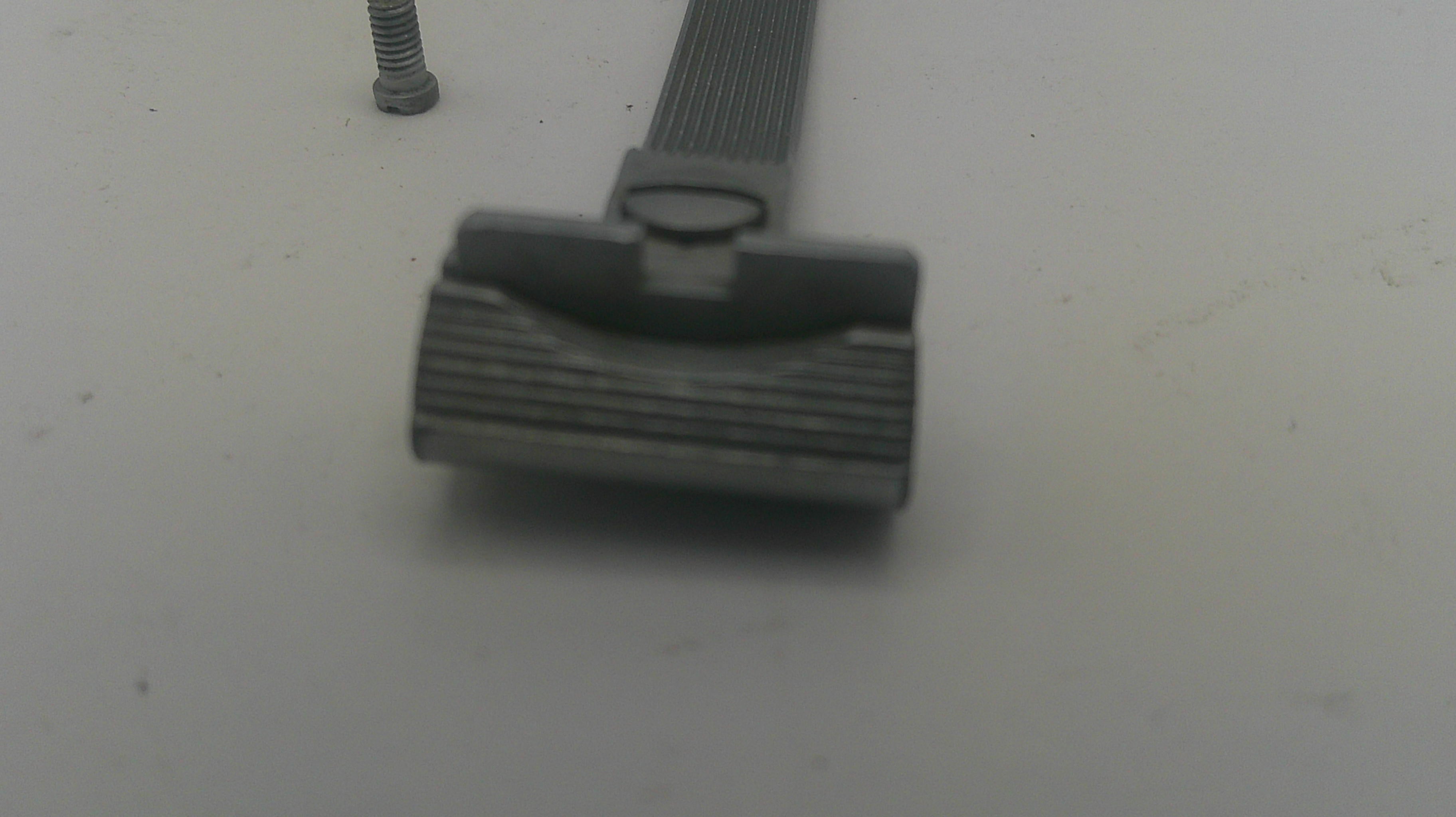 A close-up of a dark gray metal razor head with a textured guard, shown on a plain light gray surface with a small metal screw nearby.