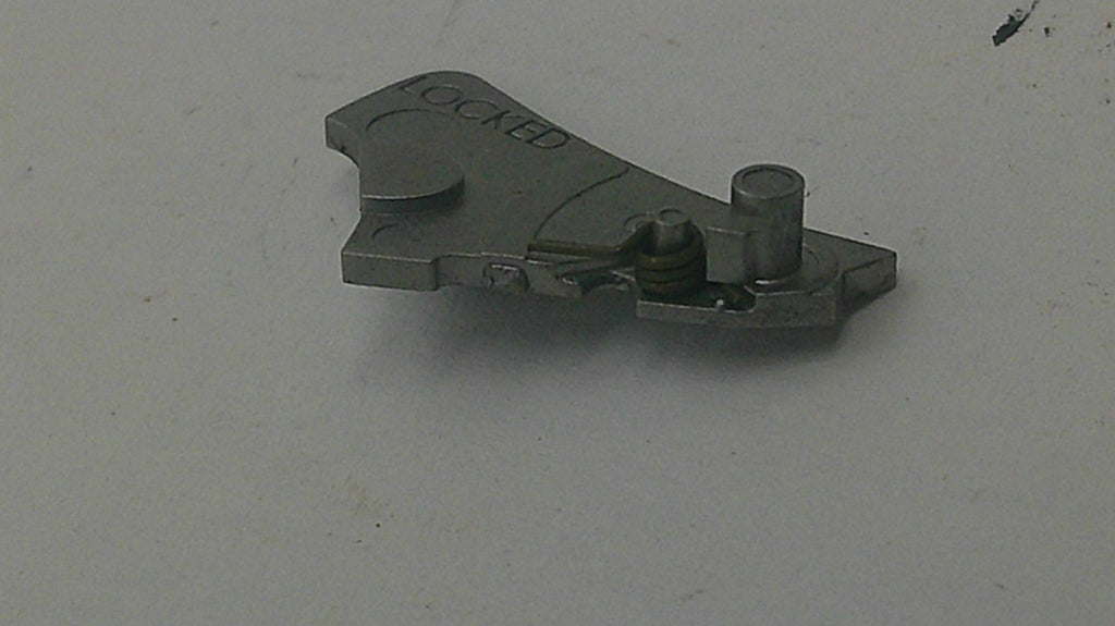 A small metallic mechanical part with the word 'LOCKED' engraved on it, photographed on a plain light gray surface.