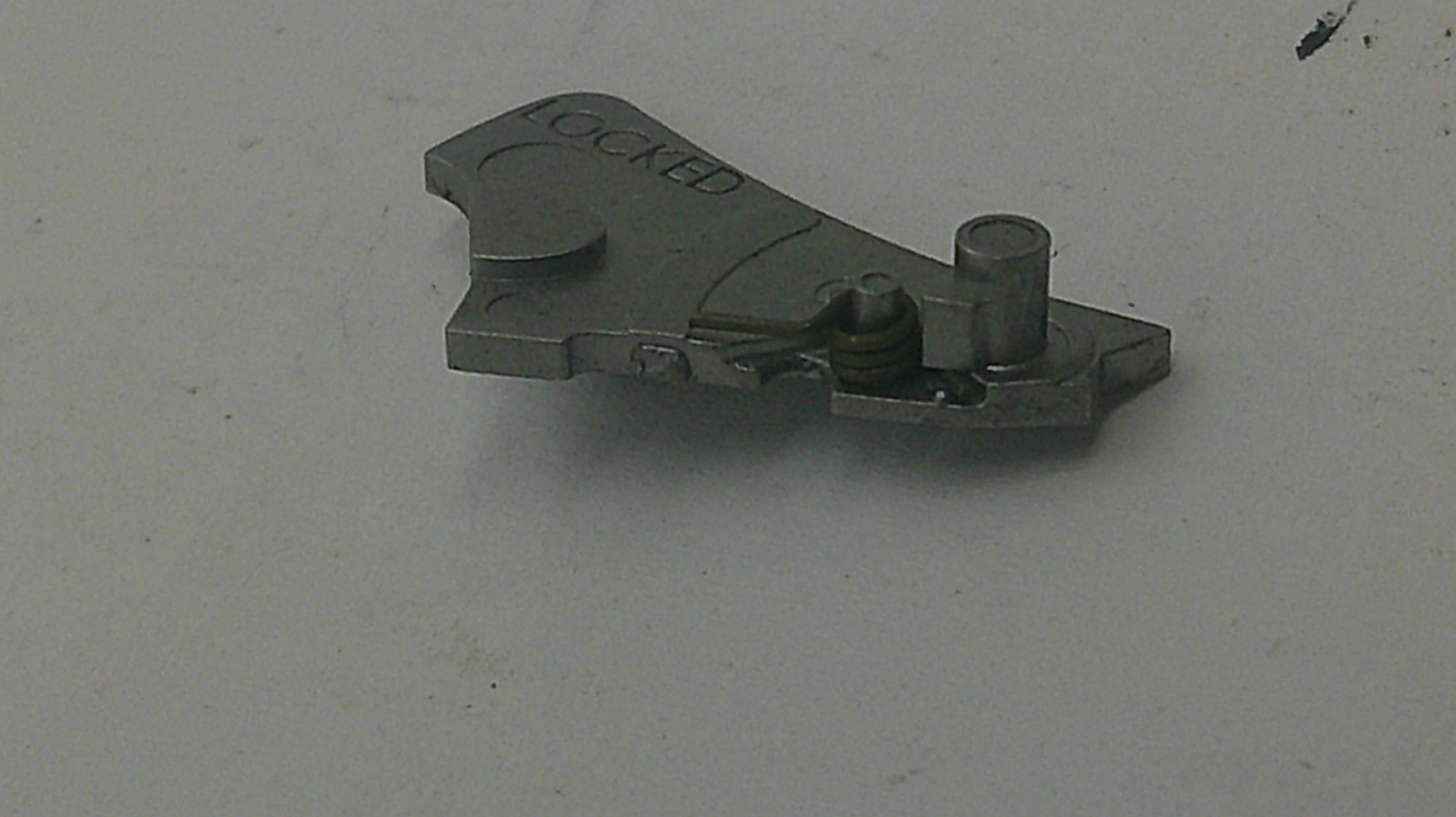 A small metallic mechanical part with the word 'LOCKED' engraved on it, photographed on a plain light gray surface.