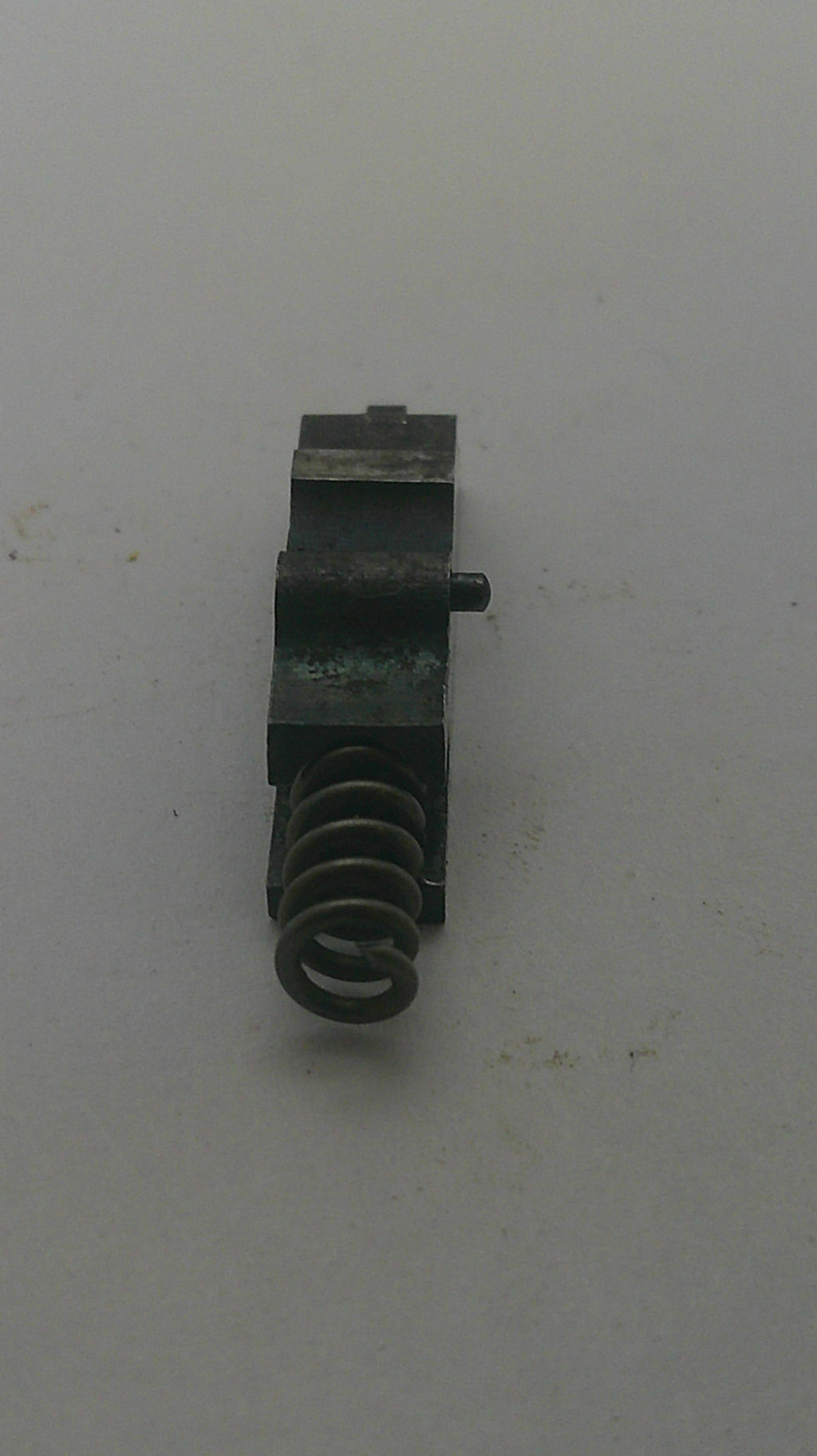 A small black metal mechanical component with a coiled spring, photographed on a plain off-white surface.