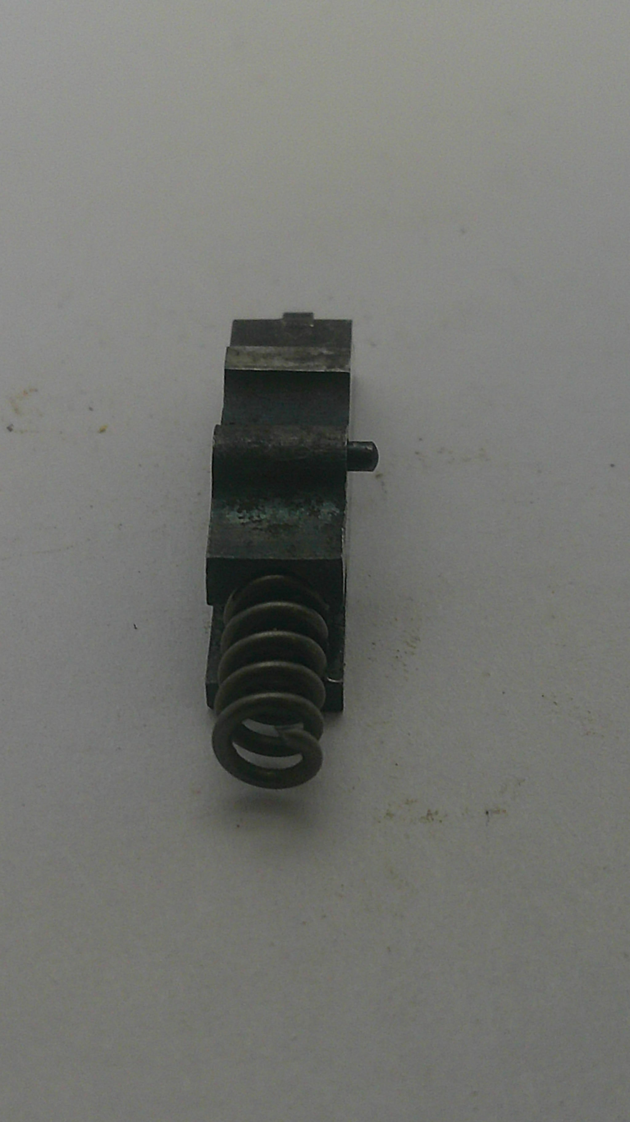 A small black metal mechanical component with a coiled spring, photographed on a plain off-white surface.