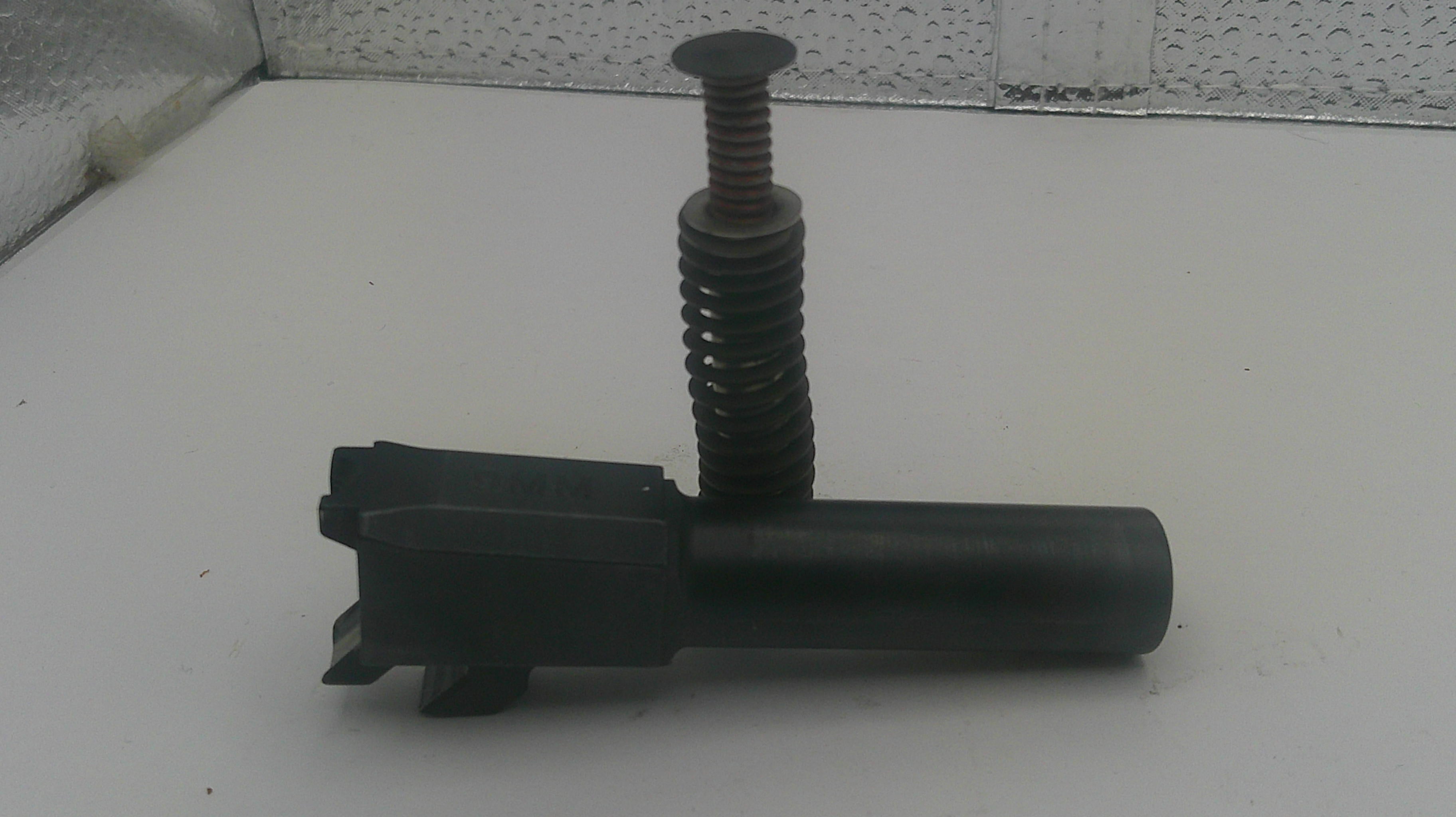 A black metal cylindrical component with a threaded vertical rod and a flat circular top, photographed on a white surface against a textured gray background.