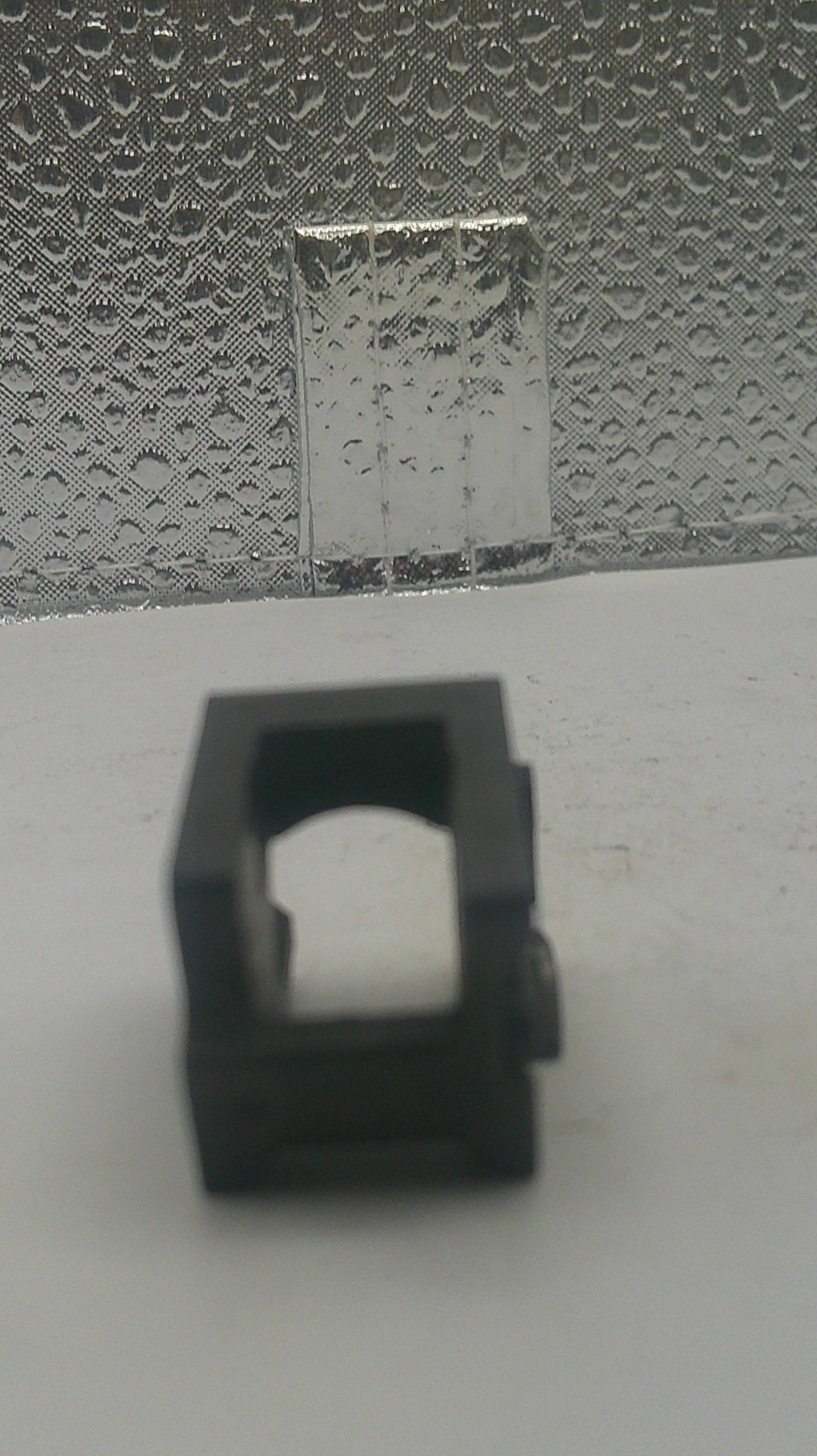 A small black plastic square-shaped clip or bracket with a hollow center, shown against a split background of plain white on the left and textured gray on the right.