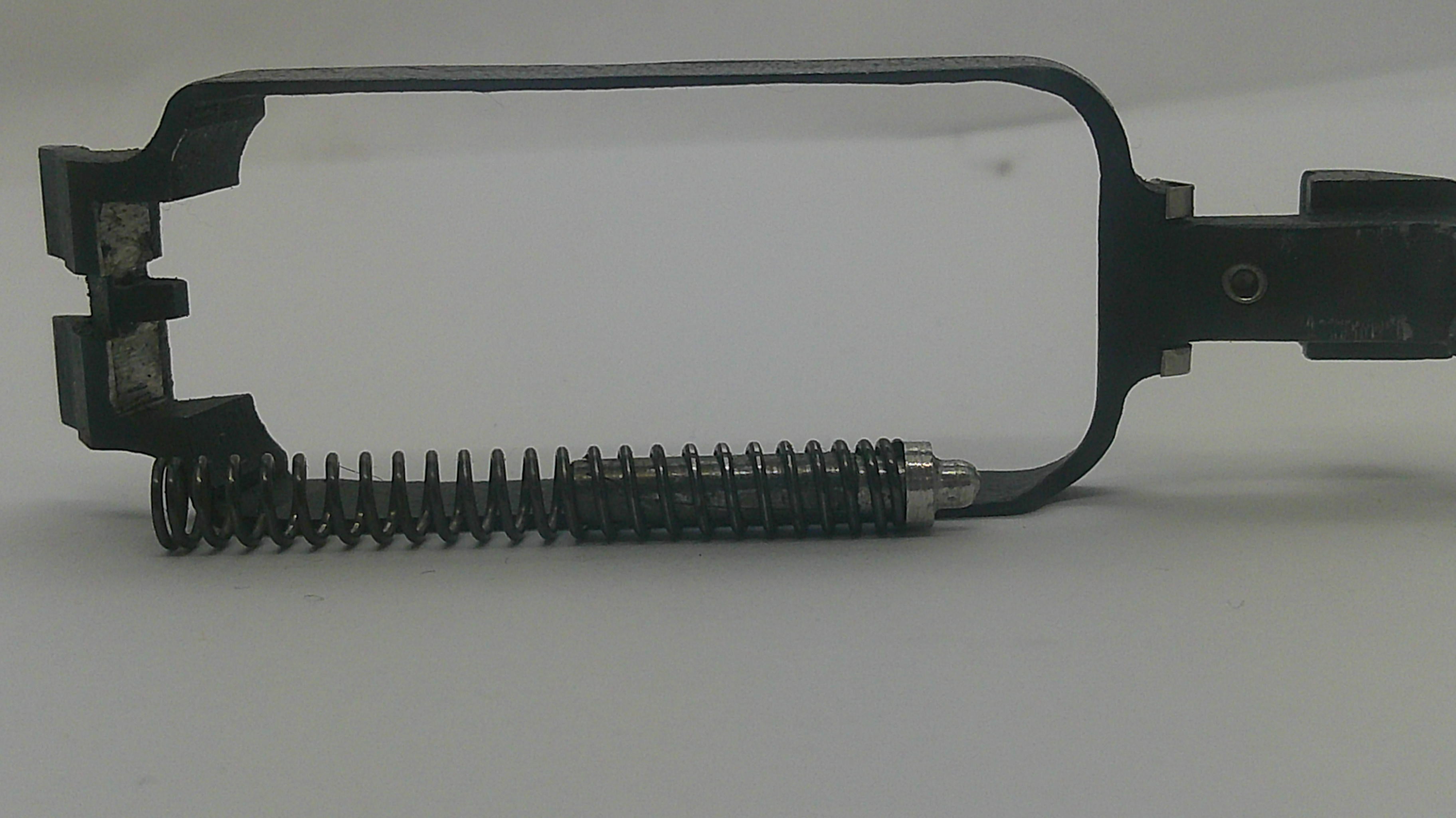 A close-up of a black metal mechanical component with a coiled spring, photographed on a plain light background.