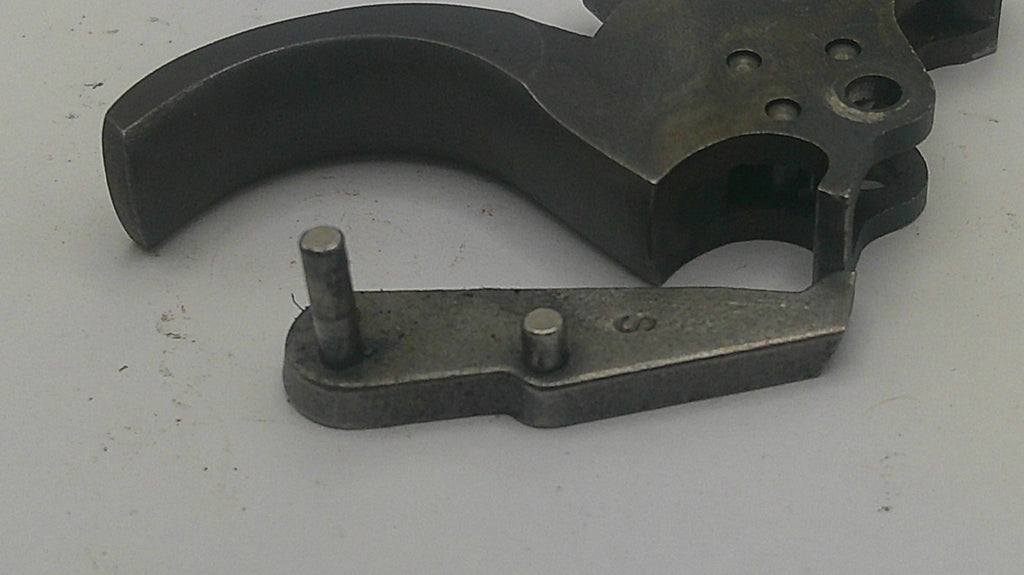 A close-up of a dark metal firearm trigger assembly component with a curved trigger and a separate flat mounting piece with two pins, shown on a light gray background.