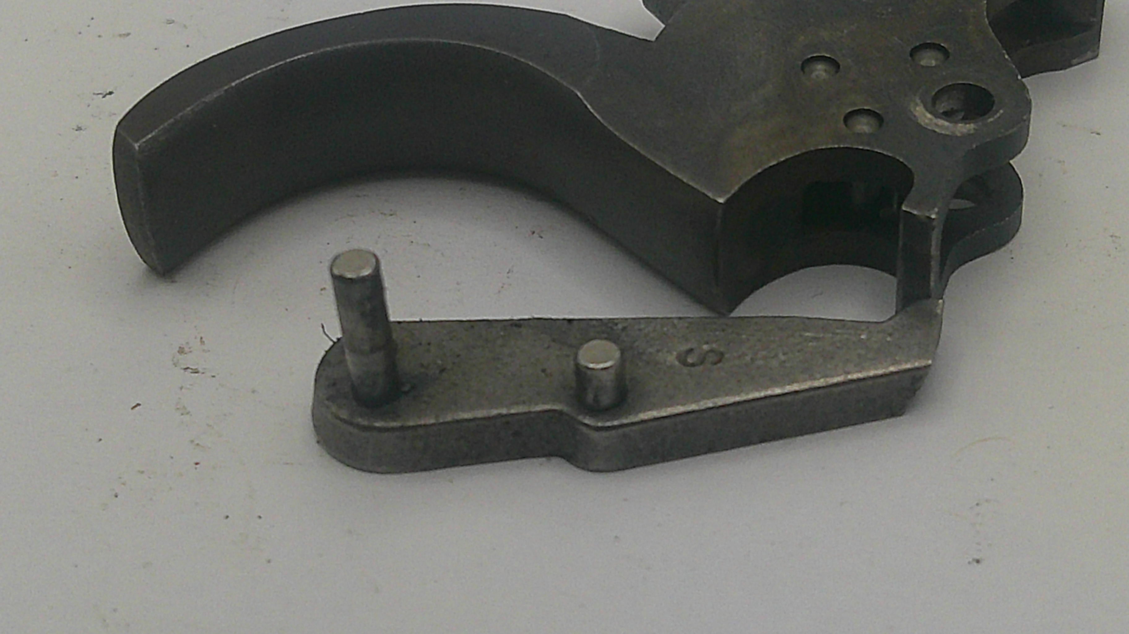 A close-up of a dark metal firearm trigger assembly component with a curved trigger and a separate flat mounting piece with two pins, shown on a light gray background.