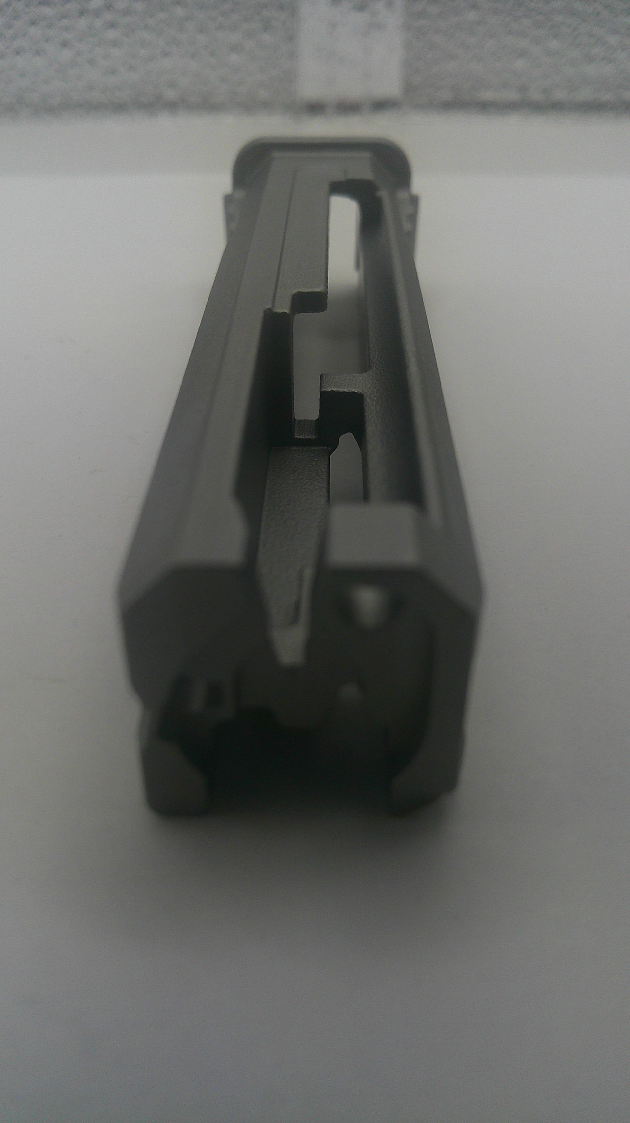 A dark gray plastic replacement part, possibly a handle grip or mechanical component, shown on a plain light gray surface.
