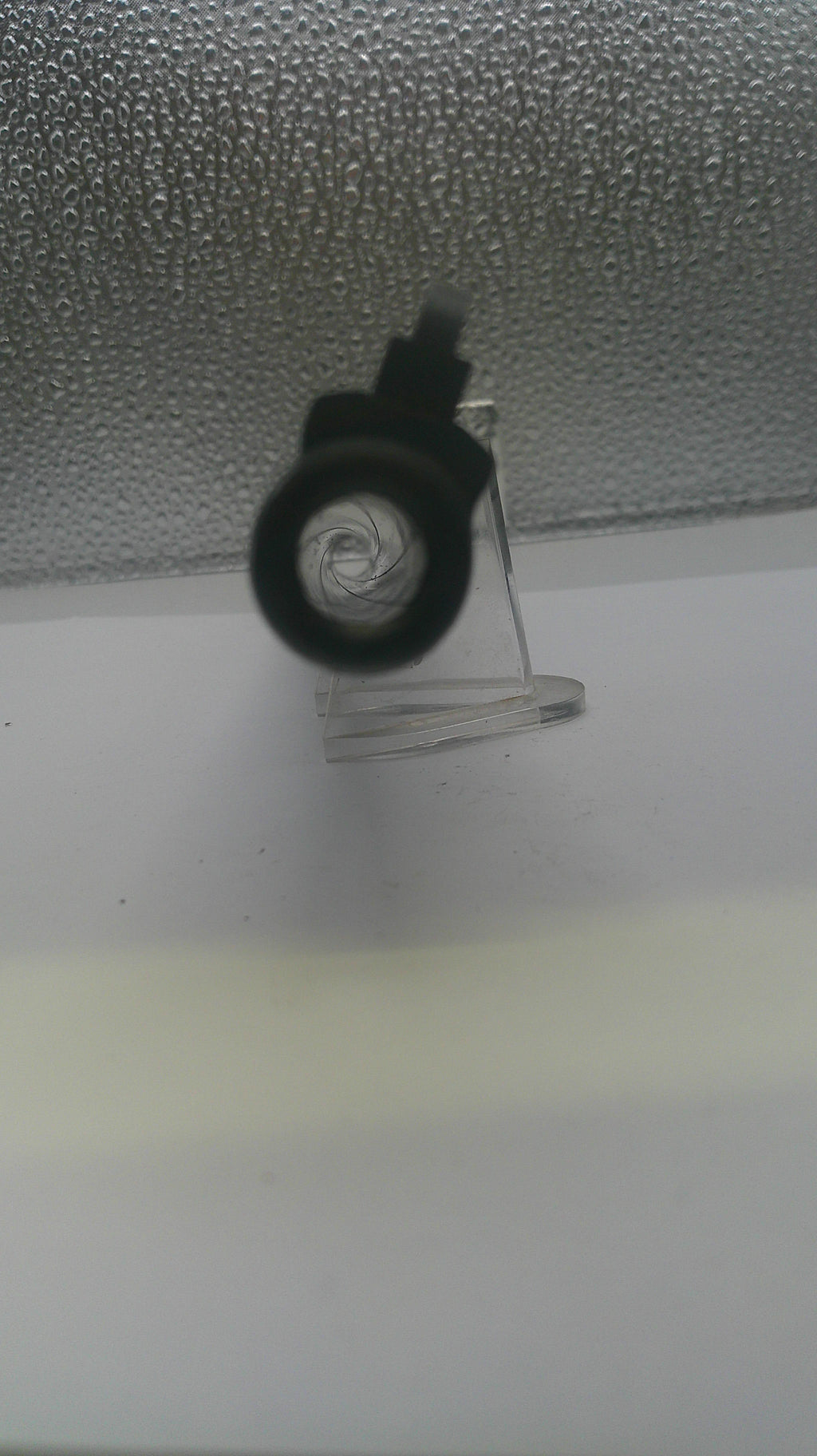 A close-up of a black circular object, possibly a lens or cap, attached to a clear stand against a textured gray and white background.