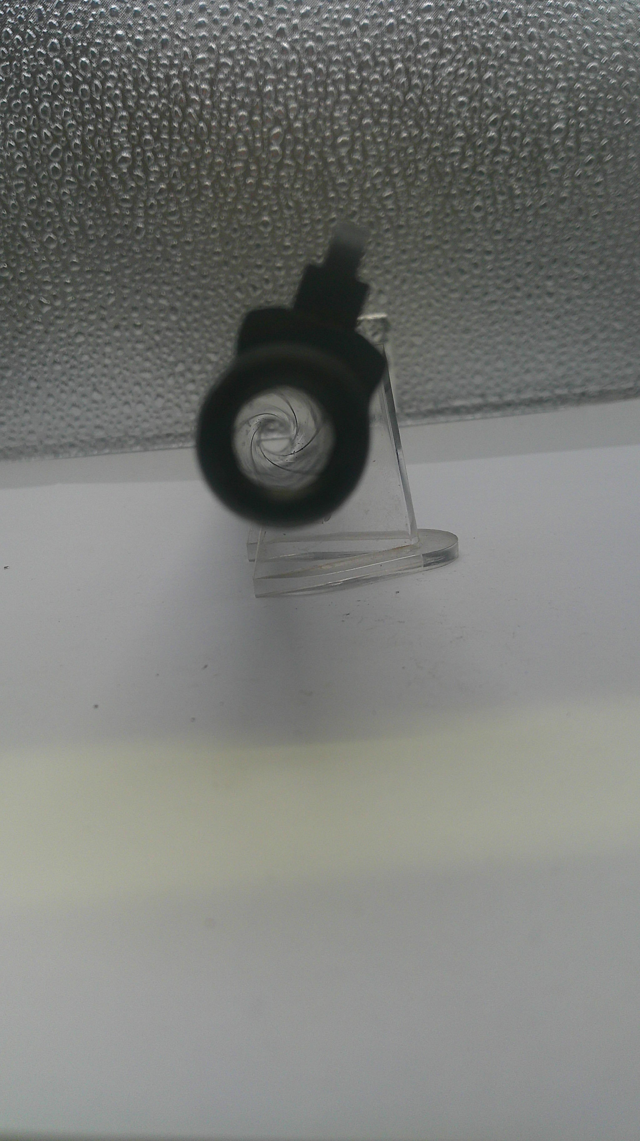 A close-up of a black circular object, possibly a lens or cap, attached to a clear stand against a textured gray and white background.