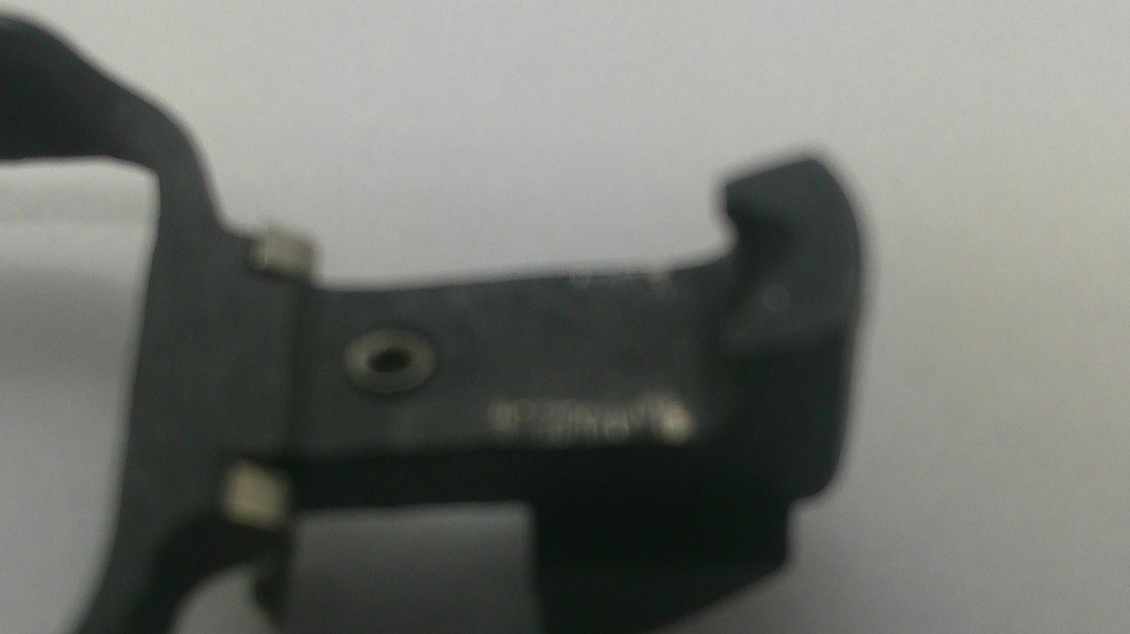 A close-up of a black plastic or metal mechanical component with a small circular hole and faint stamped markings, photographed against a plain light background.