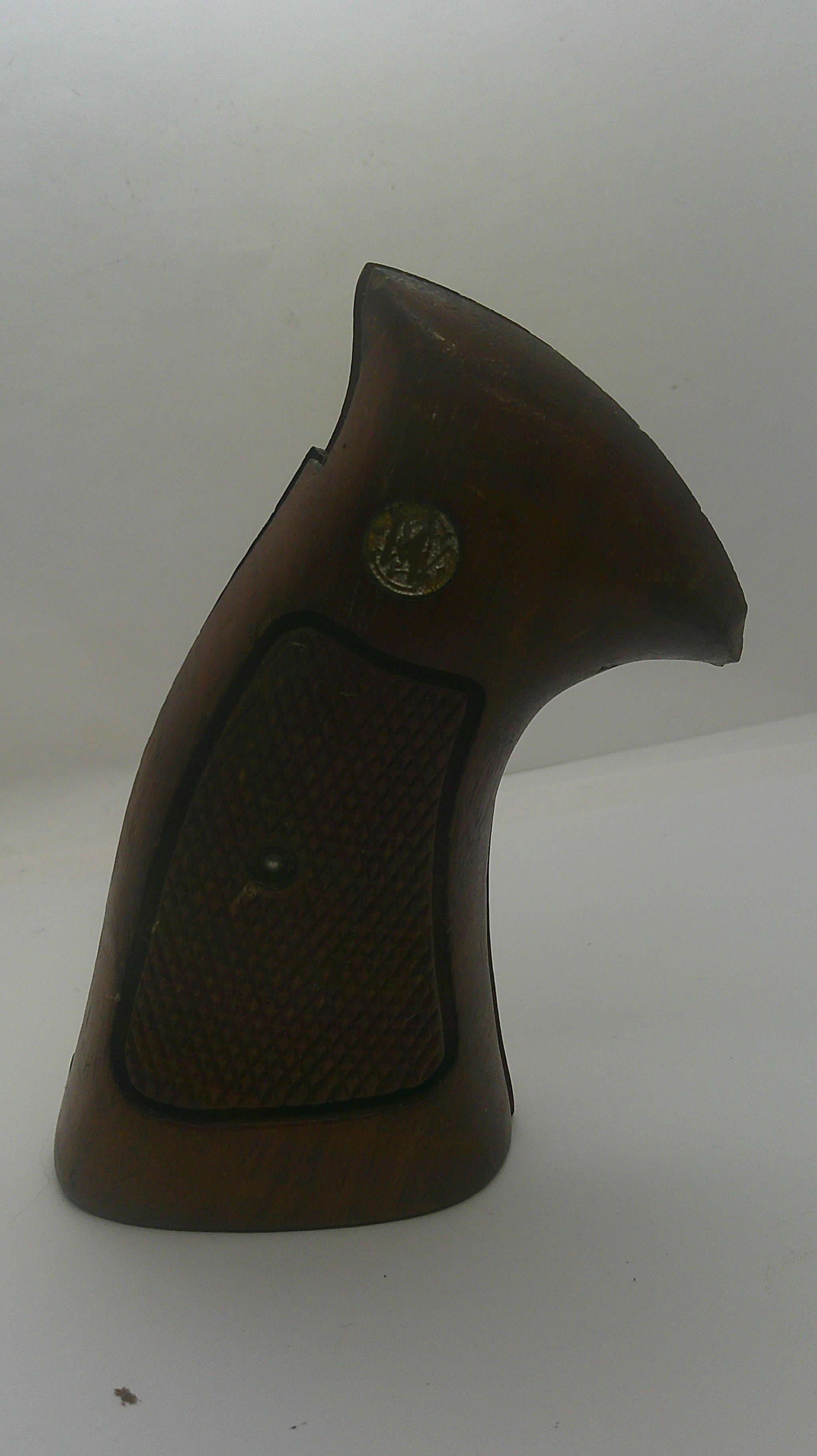A brown wooden gun stock or grip component with a textured grip panel and a small circular brass emblem, shown against a plain off-white background.