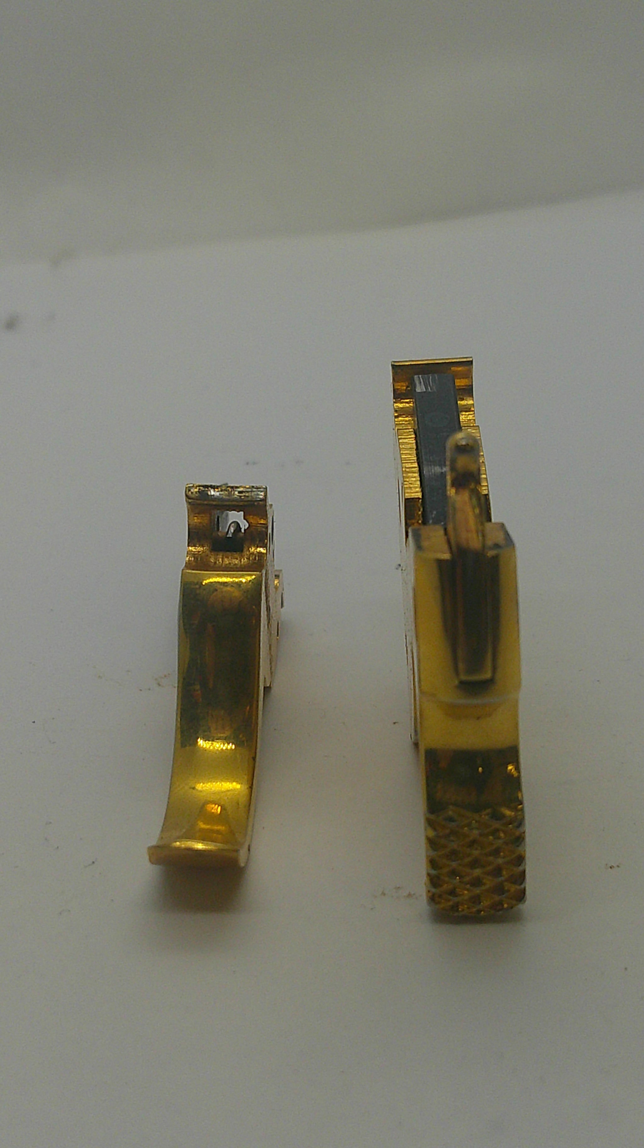 Two small gold-toned metal clips or fasteners placed vertically on a plain off-white surface.