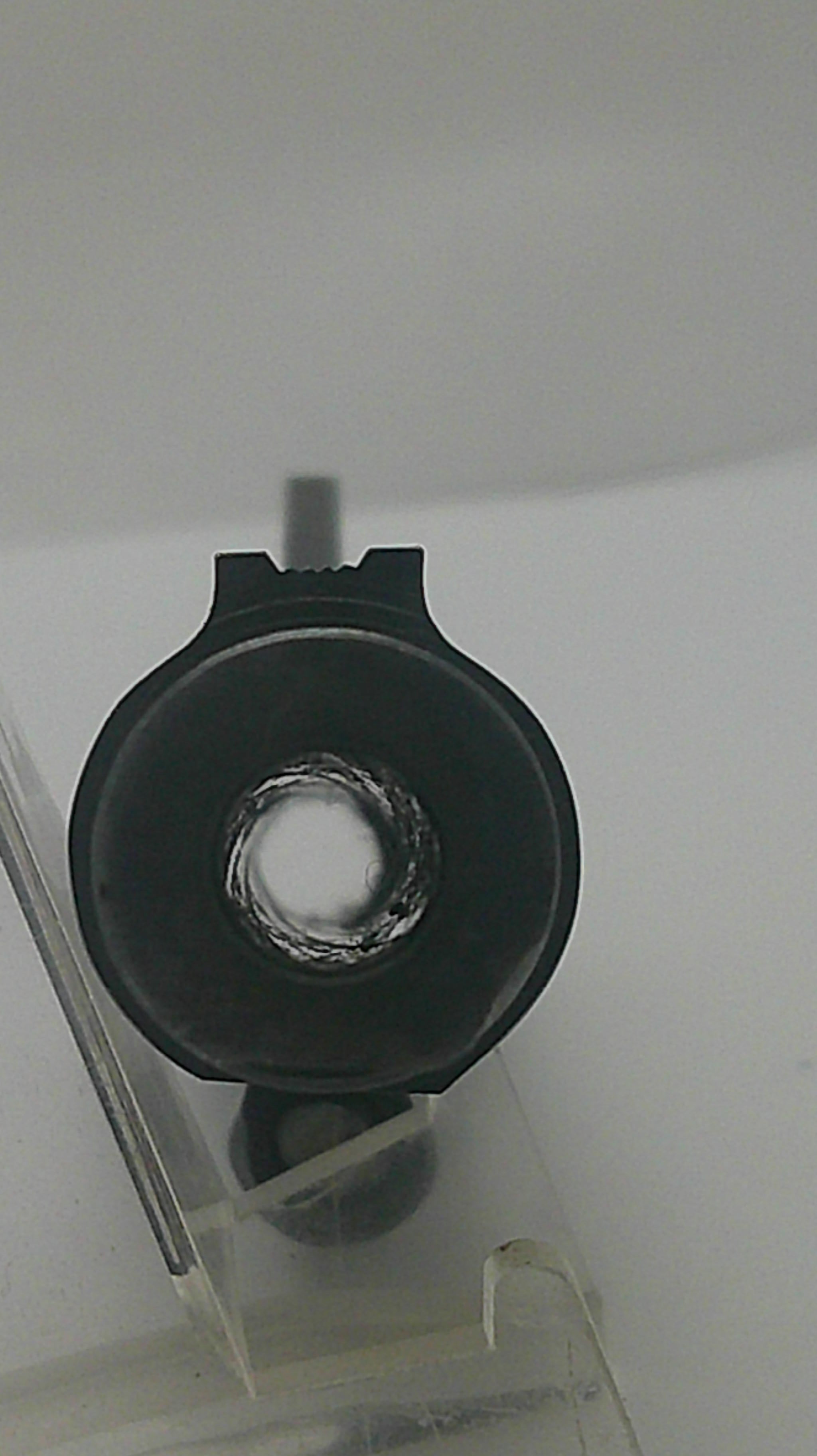 A close-up of a black circular component with a central hole, mounted on a clear plastic frame against a plain light background.