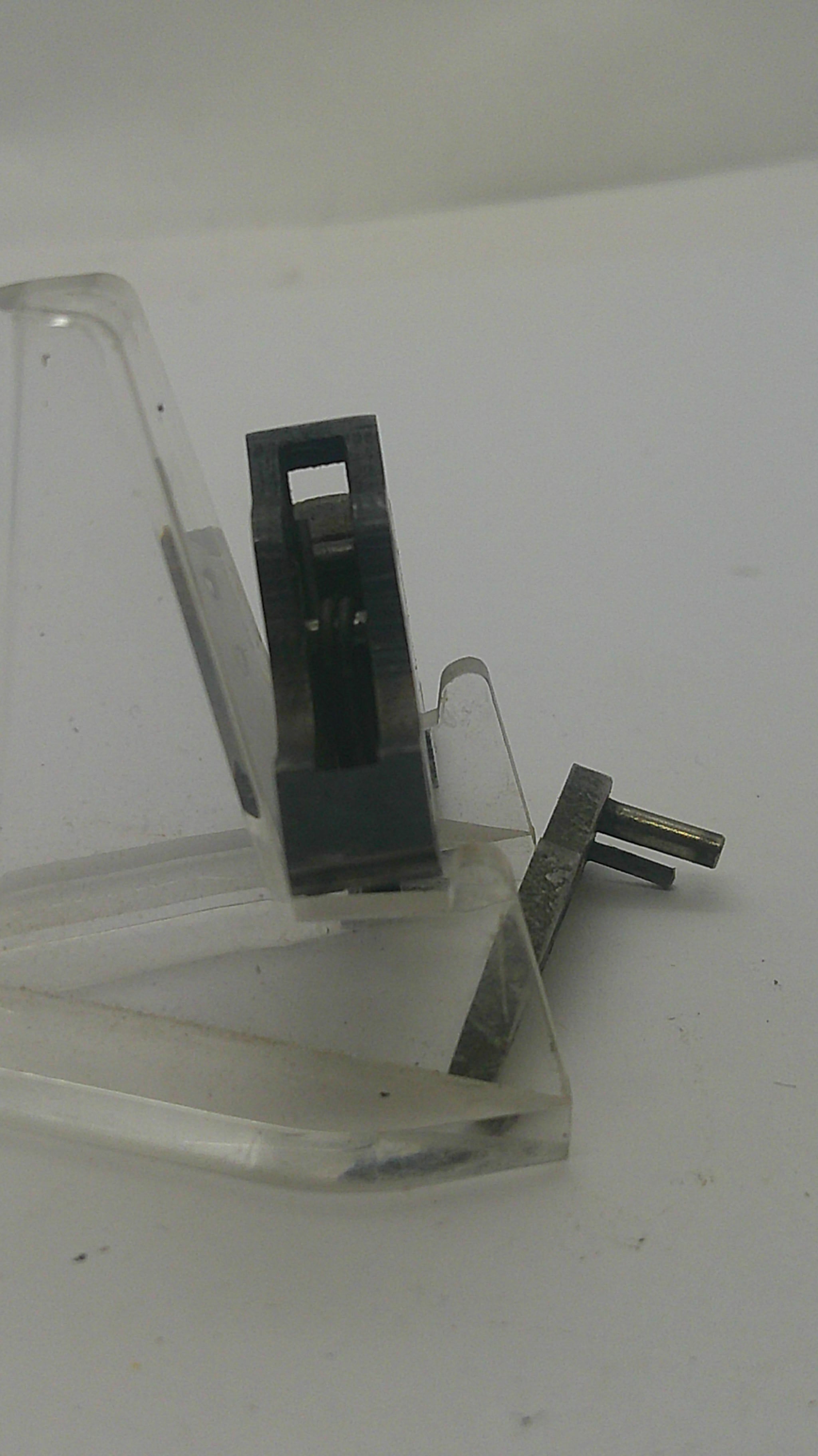 A close-up of a clear plastic mounting bracket or clip with a black metal component and two small metal pins, shown against a plain light background.