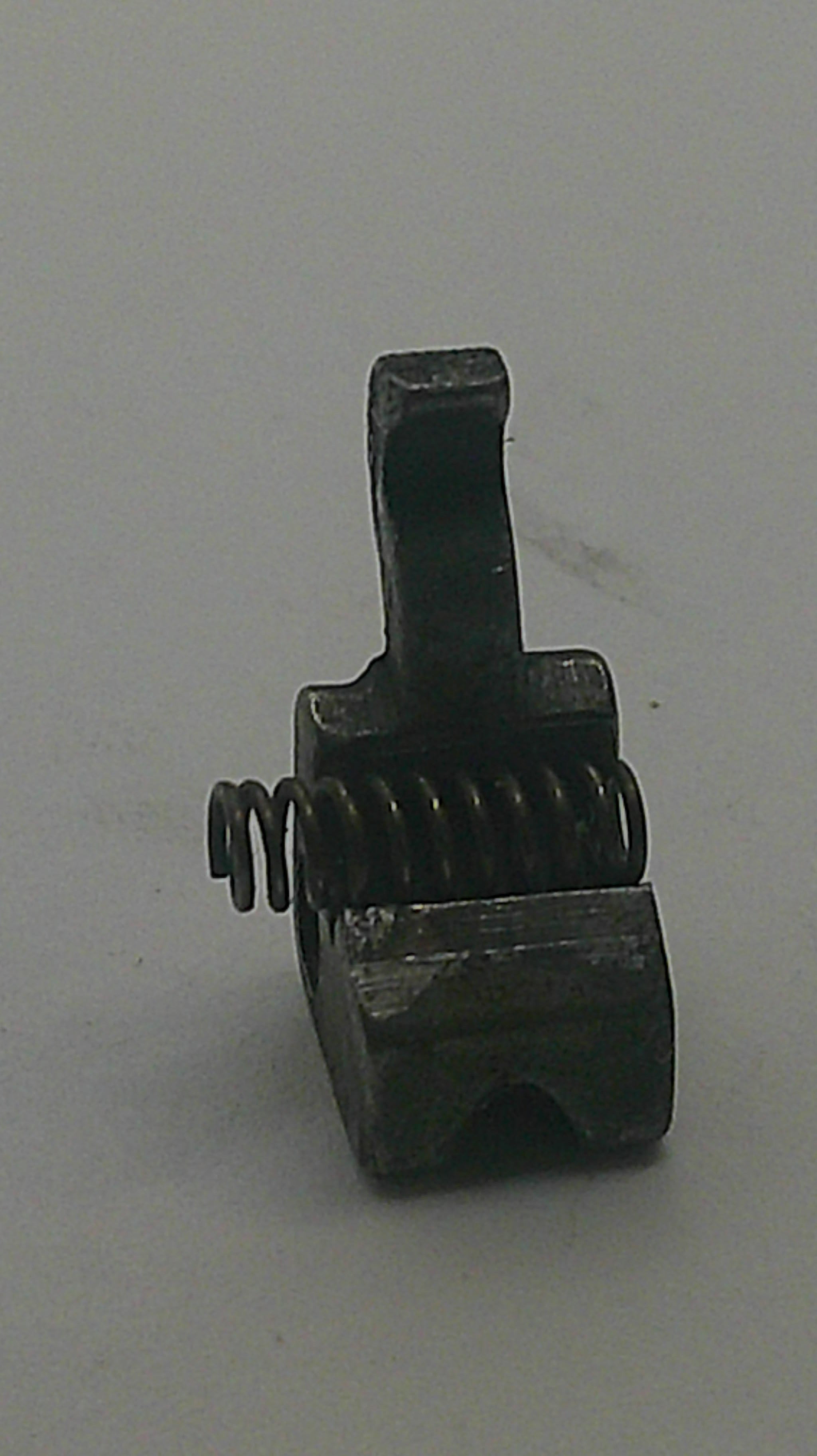 A small dark metal mechanical component with a coiled spring, photographed against a plain light gray background.