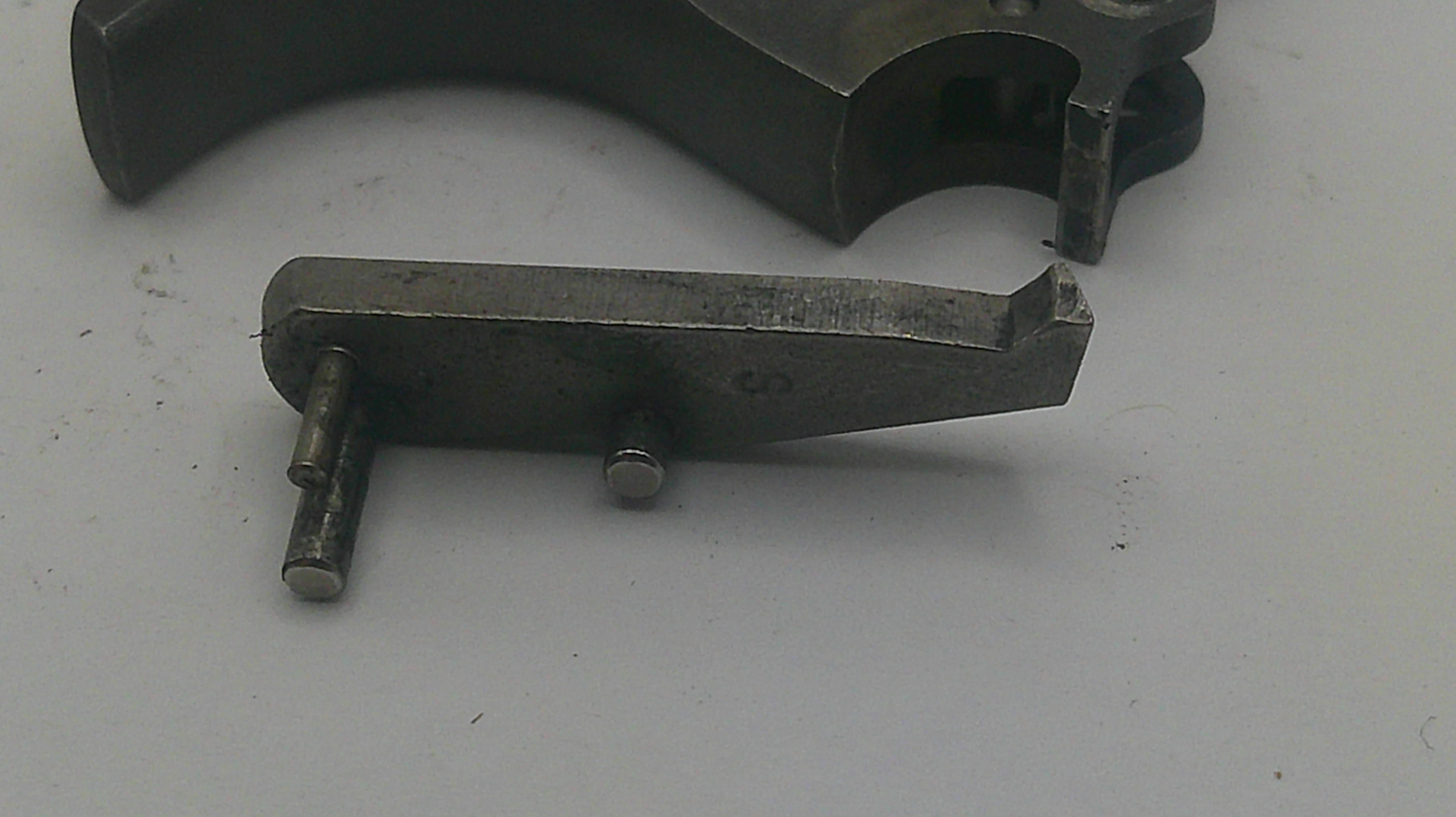 A close-up of a metallic gun trigger assembly component on a light gray surface, with another dark metal part partially visible in the upper background.