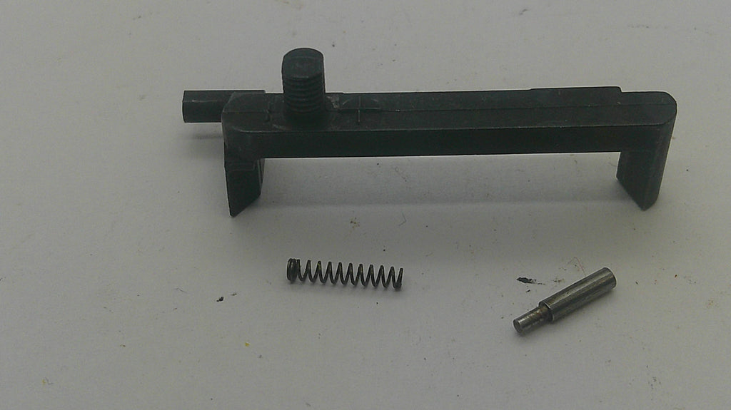 A black plastic or metal mechanical component with a small coil spring and a metal pin placed below it on a plain light background.