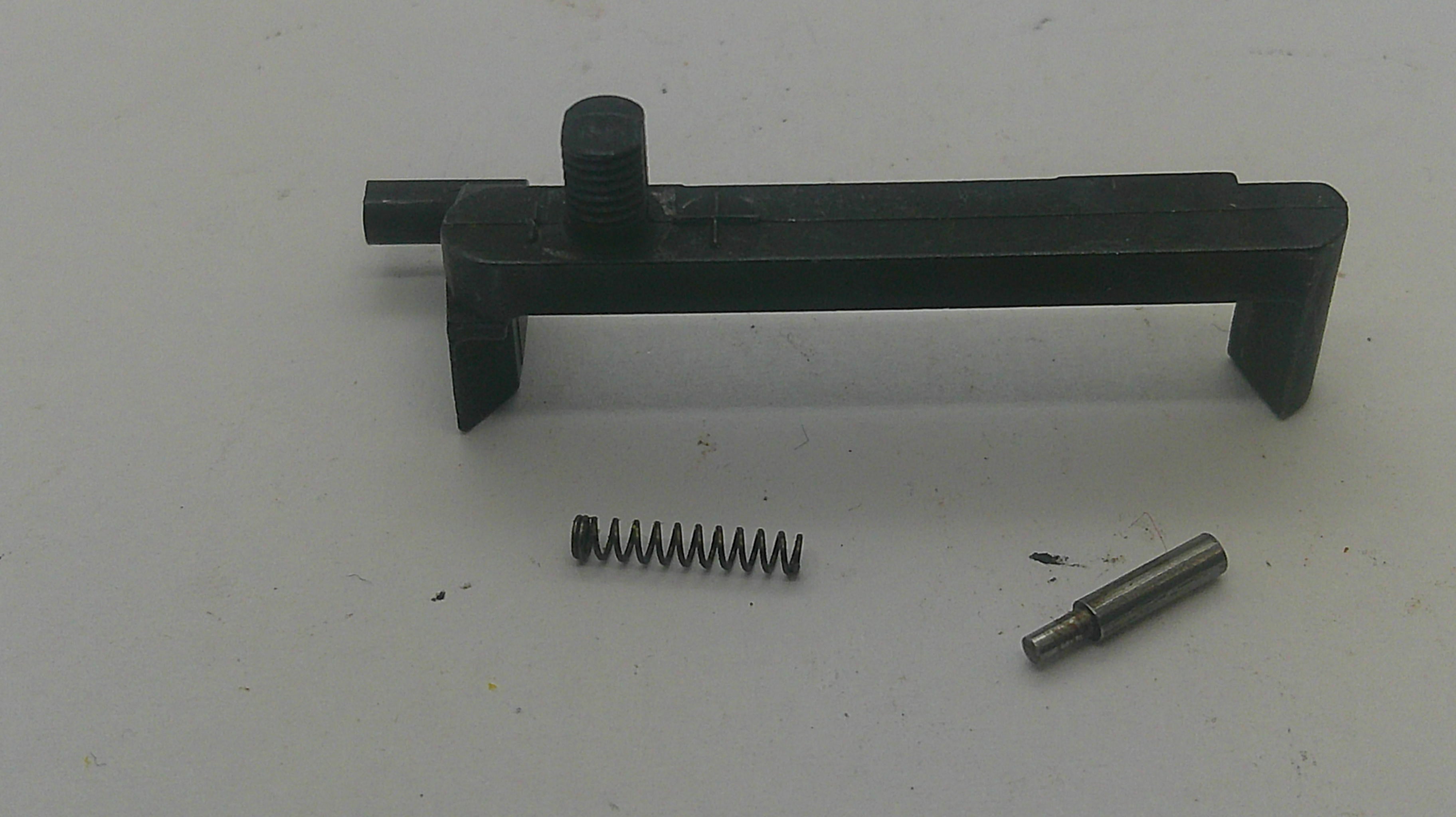 A black plastic or metal mechanical component with a small coil spring and a metal pin placed below it on a plain light background.