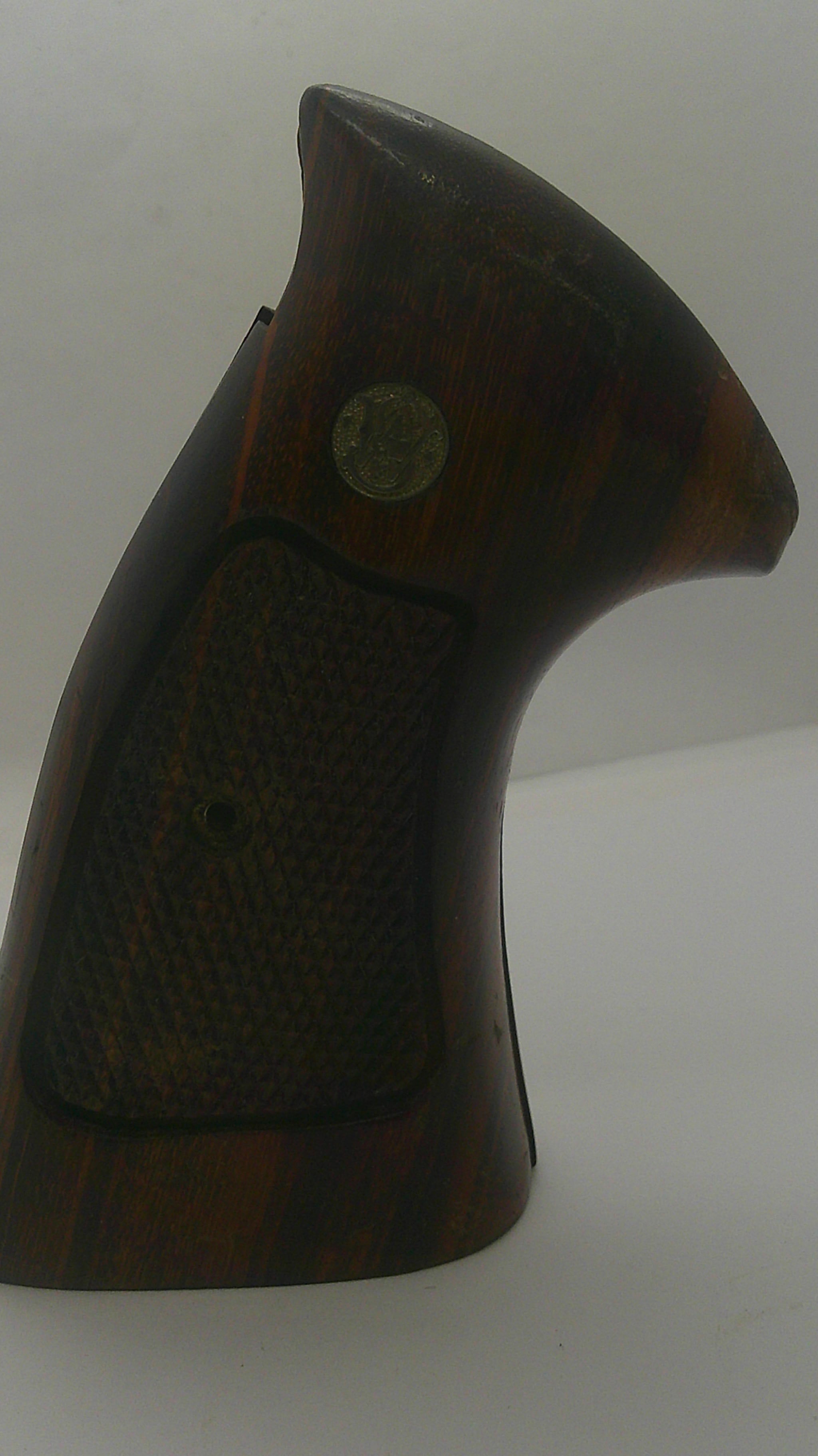 A close-up of a dark brown wooden gun stock or grip section with a textured grip panel and a small circular metal emblem, shown against a plain light background.