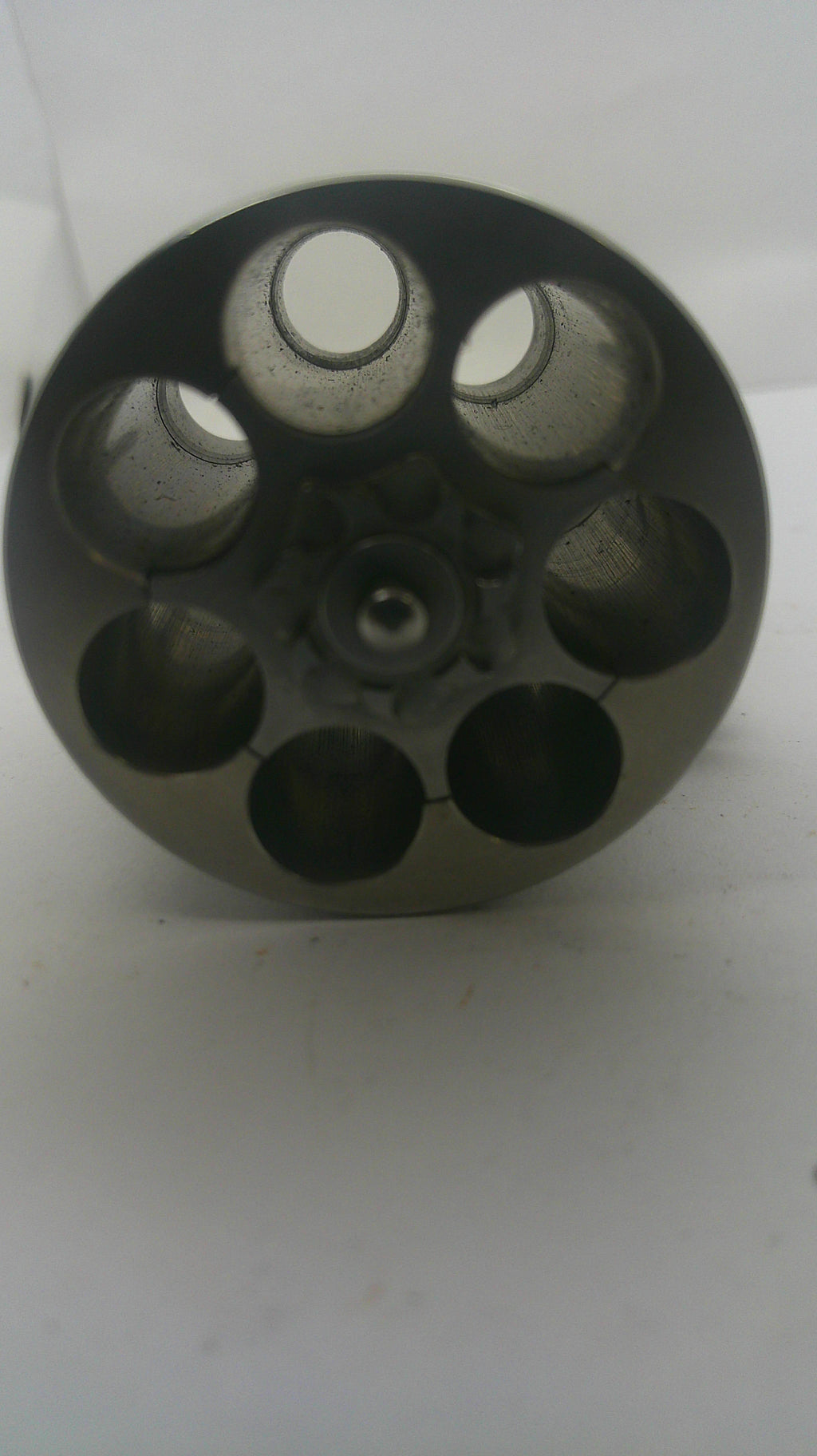 A dark gray metal revolver cylinder or magazine component with six circular chambers, photographed on a plain white background.